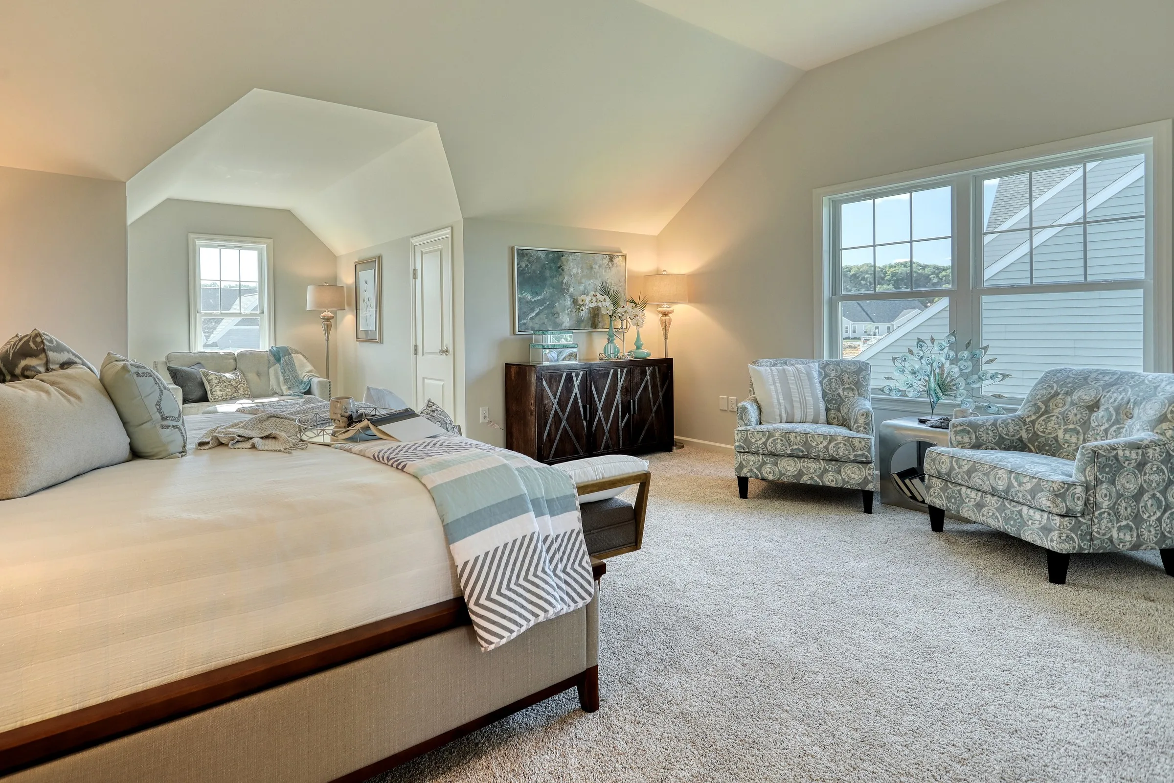 A spacious, bright bedroom with a large bed, two patterned armchairs by the window, a wooden dresser, artwork on the wall, and soft beige carpet, creating a cozy and elegant atmosphere.