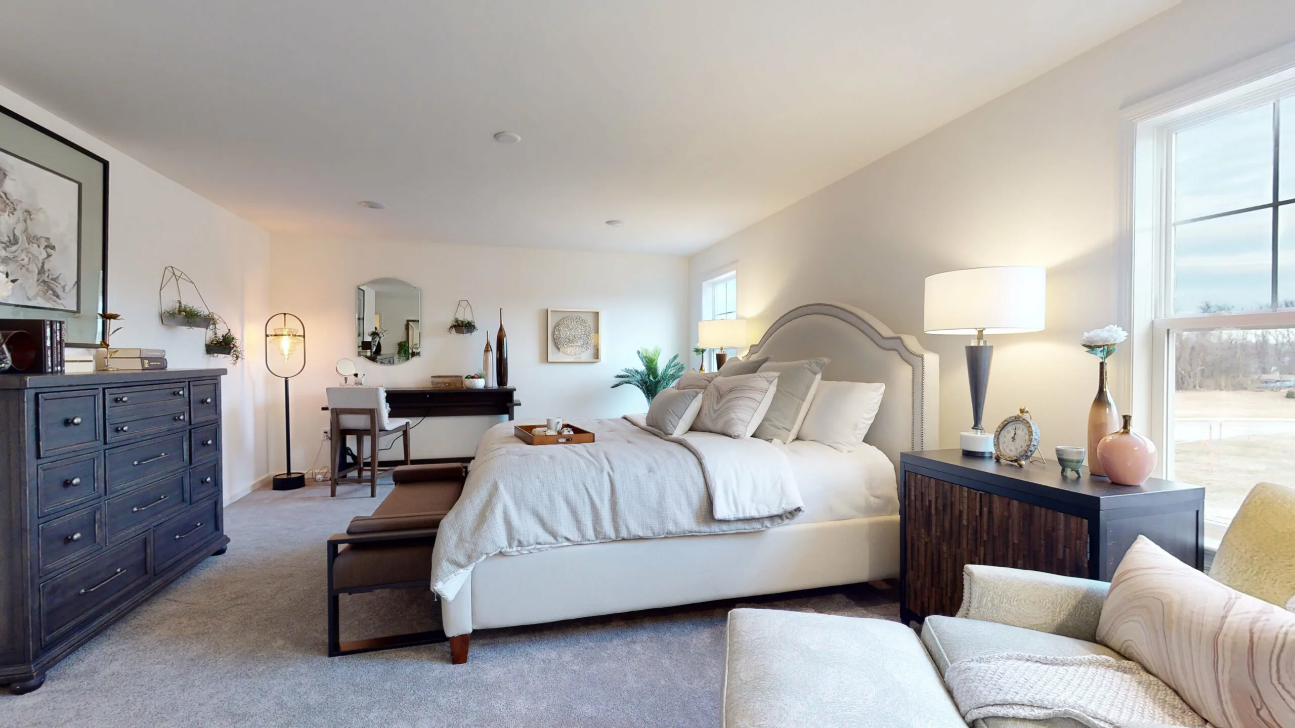 A spacious, modern bedroom with a large bed, nightstand with a lamp, dresser, armchair, writing desk, and large windows letting in natural light. Decor includes plants, framed art, and soft neutral tones.