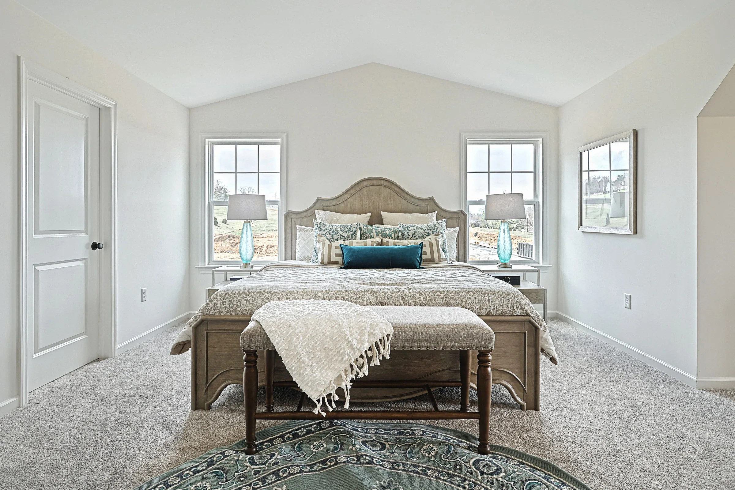 A bright, spacious bedroom with a large bed, decorative pillows, a bench at the foot, two windows, matching lamps on nightstands, and neutral, calming decor. A patterned rug lies on the carpeted floor.