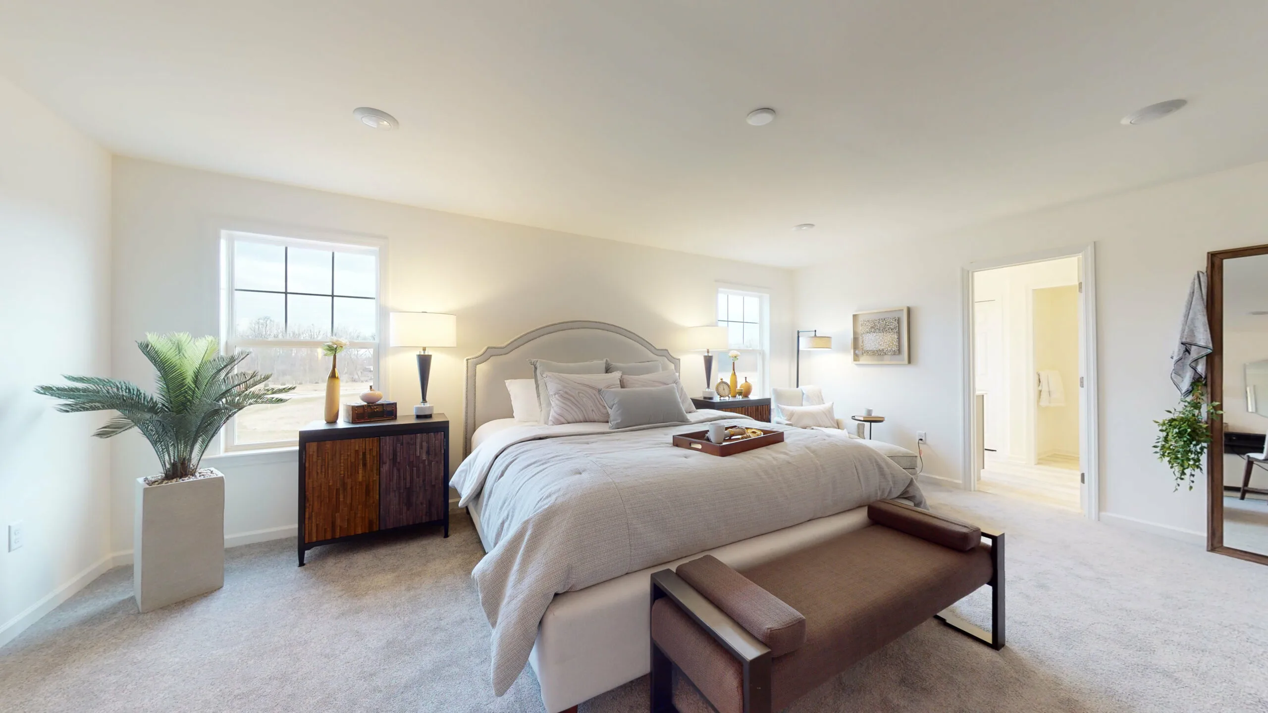 A spacious, modern bedroom with a large bed, neutral bedding, a wooden bench at the foot, two side tables with lamps, an armchair by the window, and an ensuite bathroom. Soft natural light fills the room.