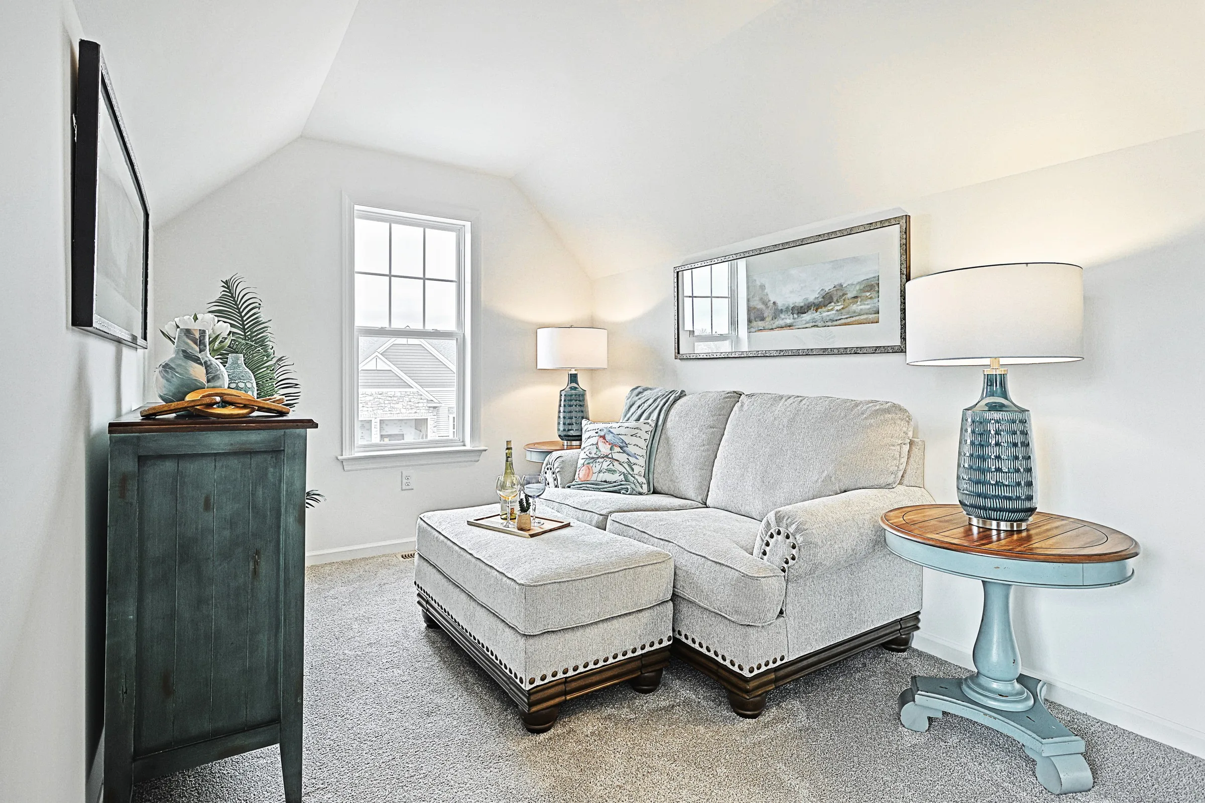 A bright, cozy living room with a light gray sofa, matching ottoman, and blue side table with a lamp. A TV is mounted on the wall, and sunlight streams through a window, illuminating the neutral decor.