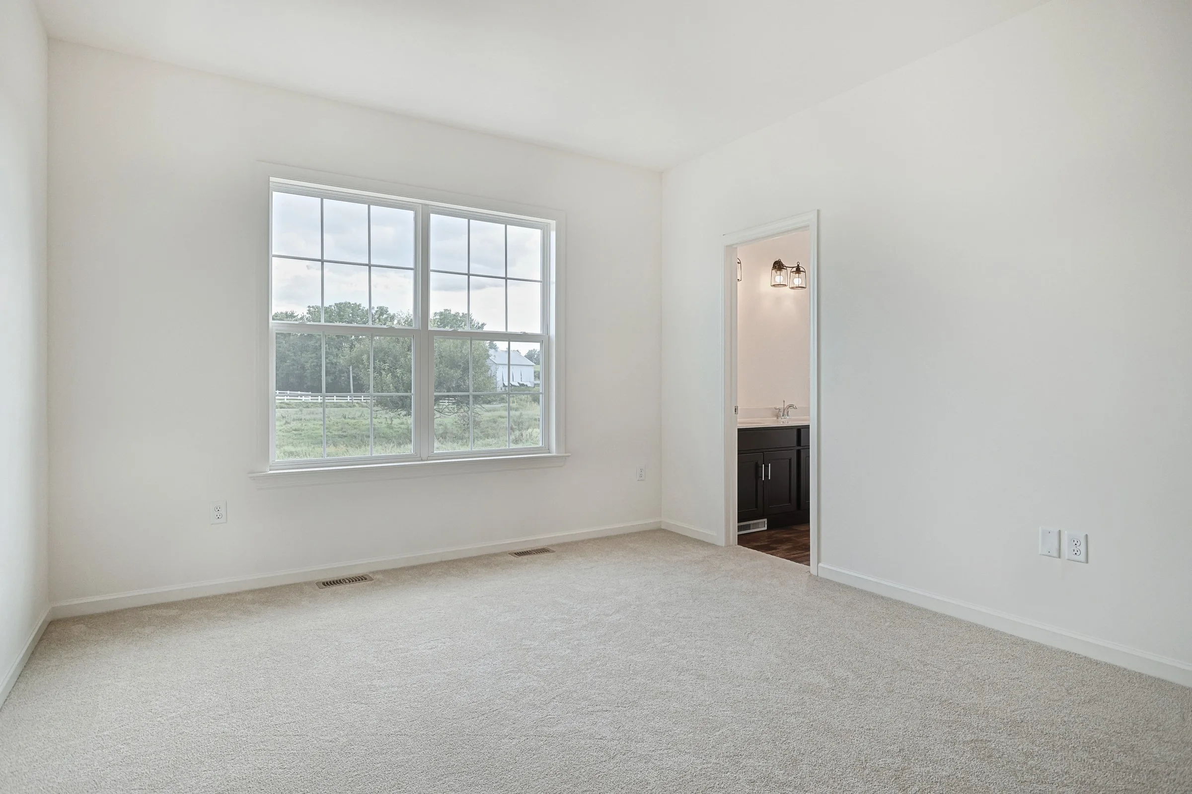 A bright, empty bedroom with beige carpet, white walls, a large window showing greenery outside, and an open doorway leading to a bathroom with dark cabinetry.