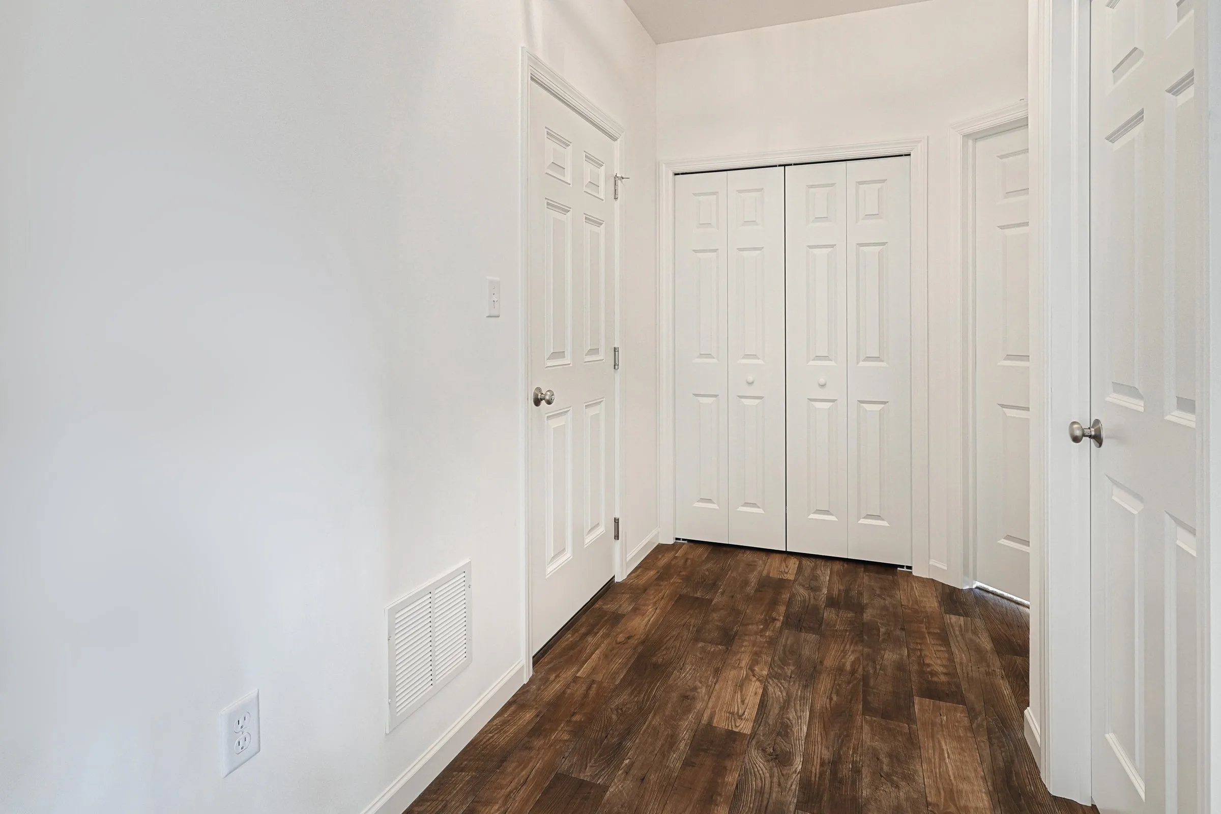 A room with white walls, three white doors, double closet doors, and wood-look flooring. The space appears empty and well-lit, with a minimal, clean design.