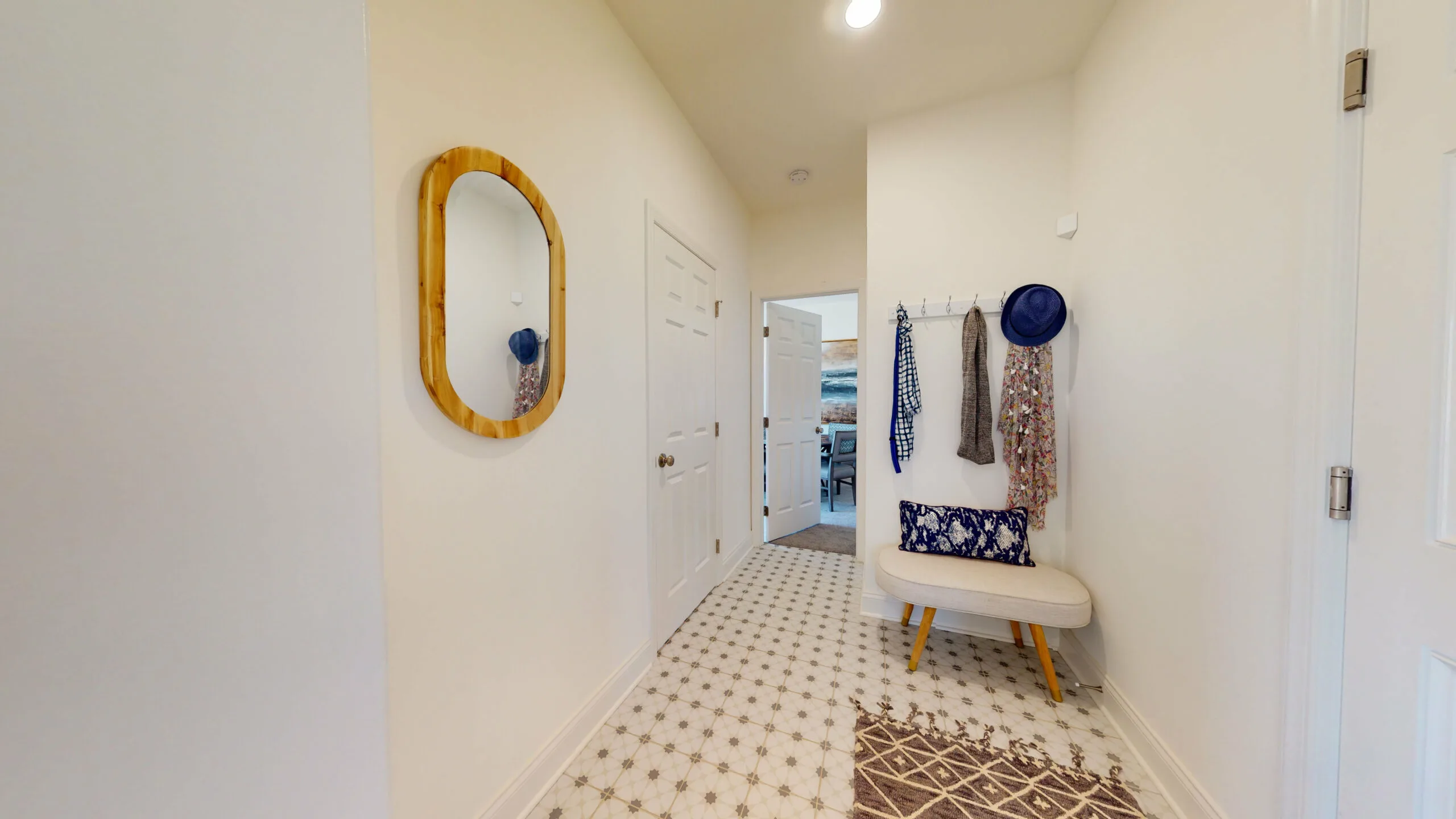 A bright hallway with patterned tile floor, a bench with blue print pillow, wall hooks holding hats and scarves, and a wooden-framed mirror on the left wall. A doorway at the end leads to another room.