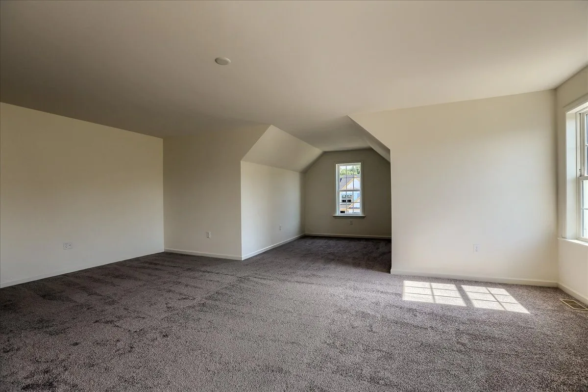 Empty room with light-colored walls, gray carpet, sloped ceilings, and a single window letting in natural light. The room is spacious with an alcove area and minimal architectural details.