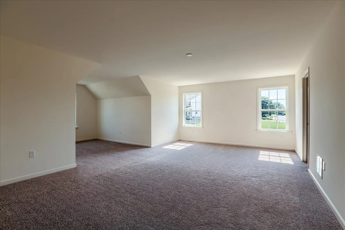 Spacious empty room with beige walls, carpeted floor, two large windows letting in natural light, and an alcove area to the left.
