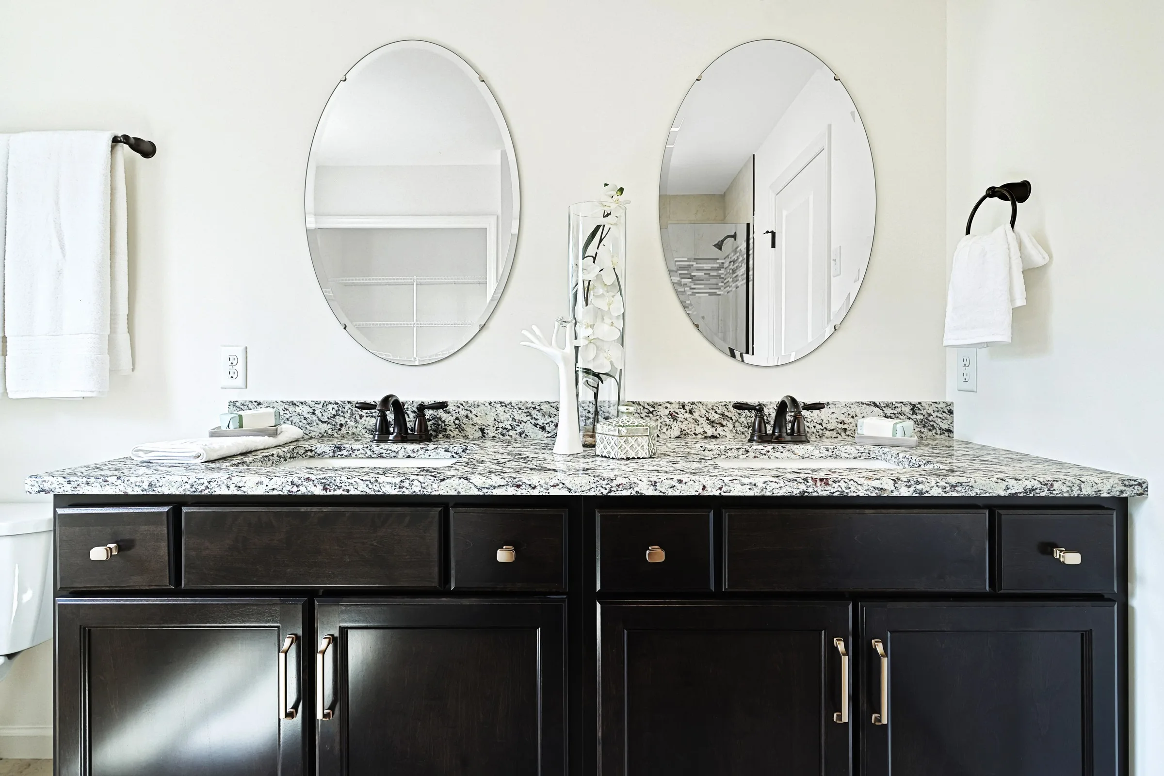 A modern bathroom with a granite countertop, two oval mirrors, dual sinks with black fixtures, dark wood cabinets, white towels, and a decorative white vase with flowers.