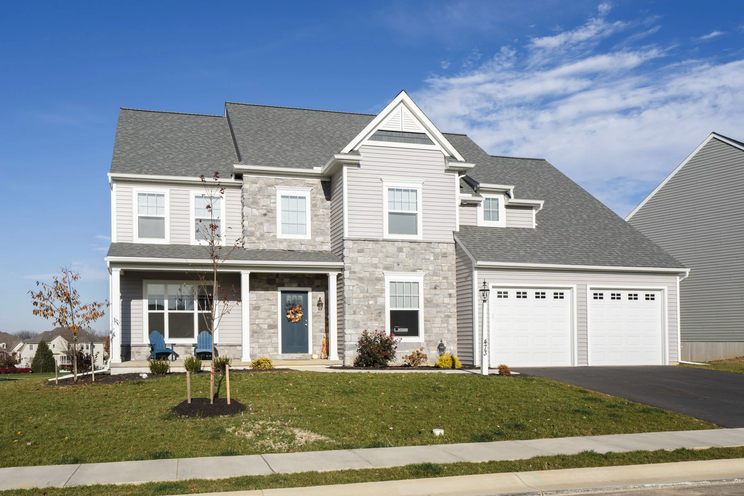 A two-story suburban house with light gray siding and stone accents, a front porch with chairs, a double garage, and a neatly manicured lawn under a blue sky with scattered clouds.