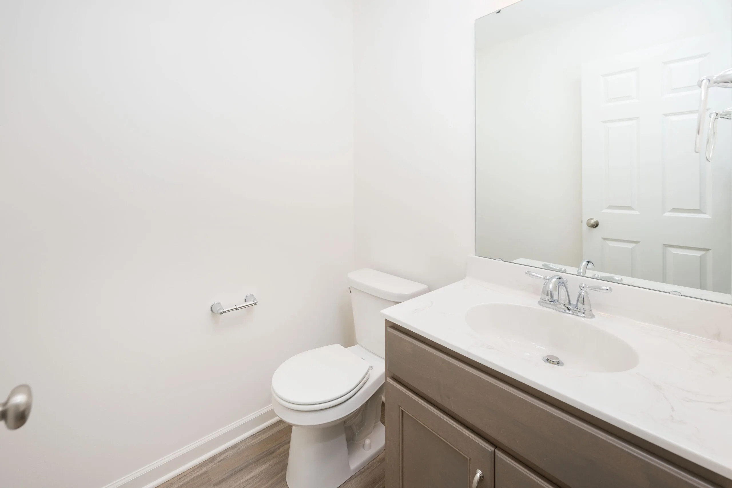 A clean, modern bathroom with a white toilet, a wall-mounted toilet paper holder, a large mirror above a sink with a brown vanity, and light wood flooring. The walls are plain white.
