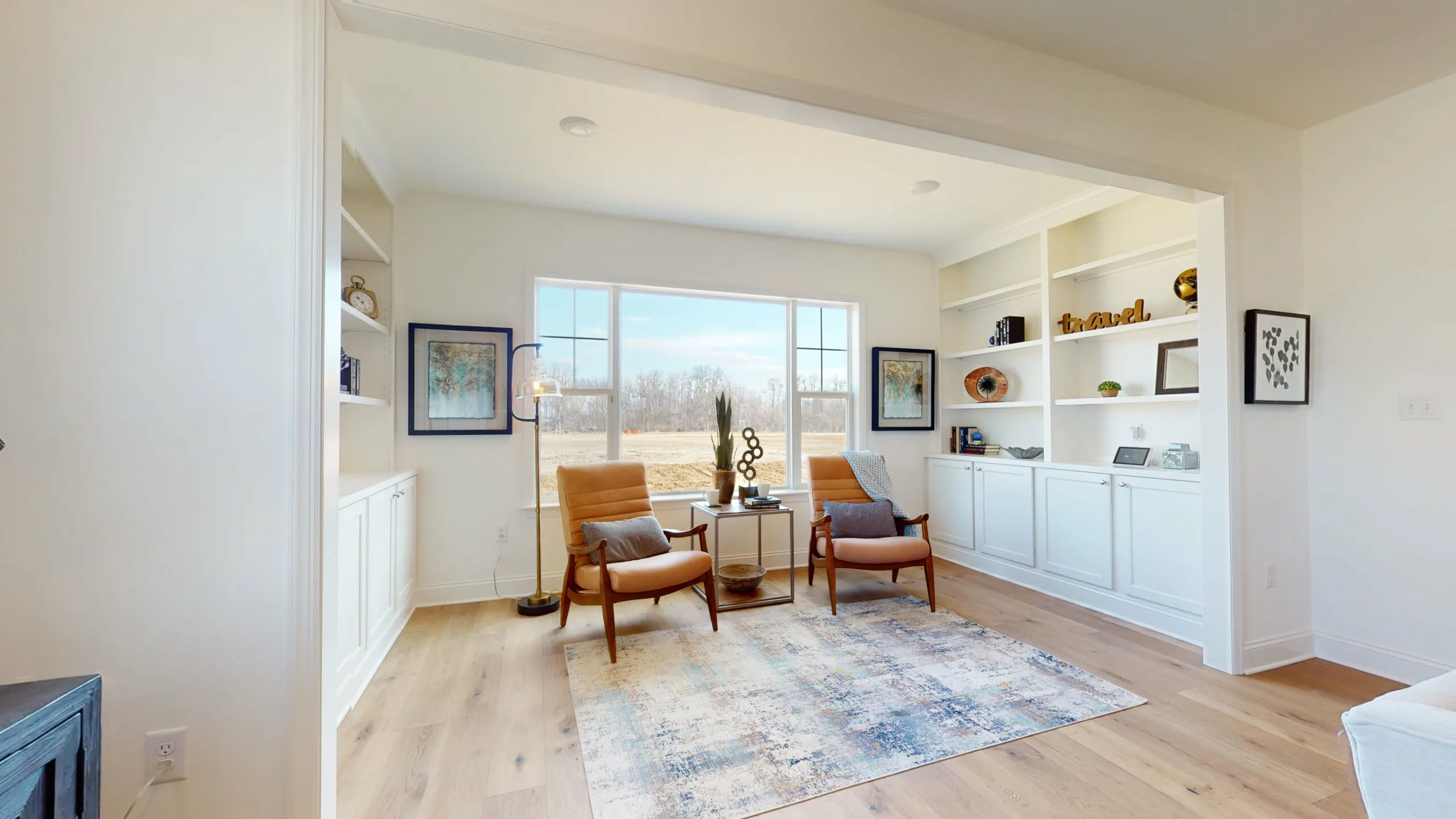 Bright sitting room with two mid-century modern chairs, a small round table, built-in white bookshelves, and large windows overlooking an outdoor landscape. A patterned rug covers the light wood floor.