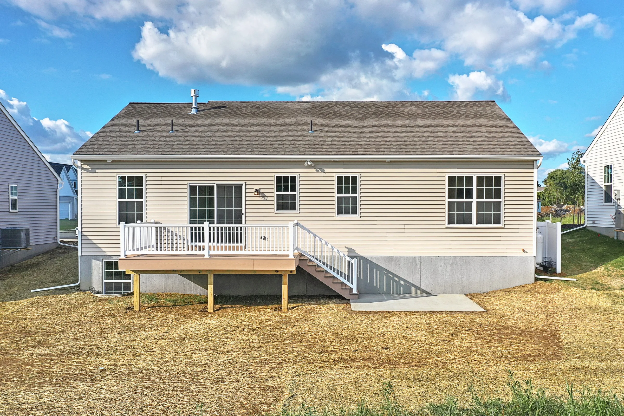 A beige single-story house with a gray roof, white-framed windows, and a small raised deck with white railings and stairs leading to a dry, straw-covered backyard under a partly cloudy sky.