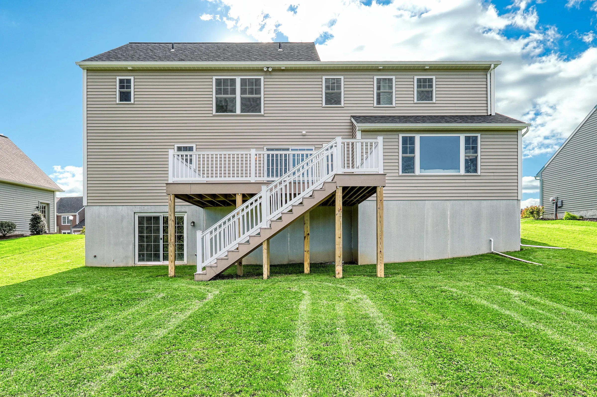 The back of a two-story house with beige siding, white-framed windows, and a raised wooden deck with stairs leading down to a large, green lawn on a sunny day.
