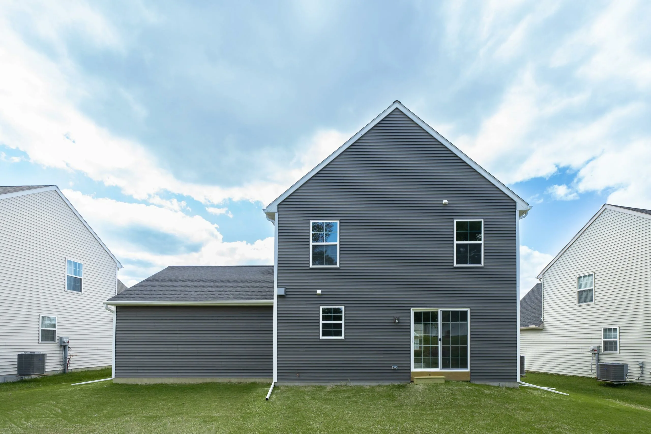 A two-story gray house with white trim, featuring sliding glass doors and several windows, sits on a grassy lawn under a partly cloudy sky. Similar houses are visible on either side.