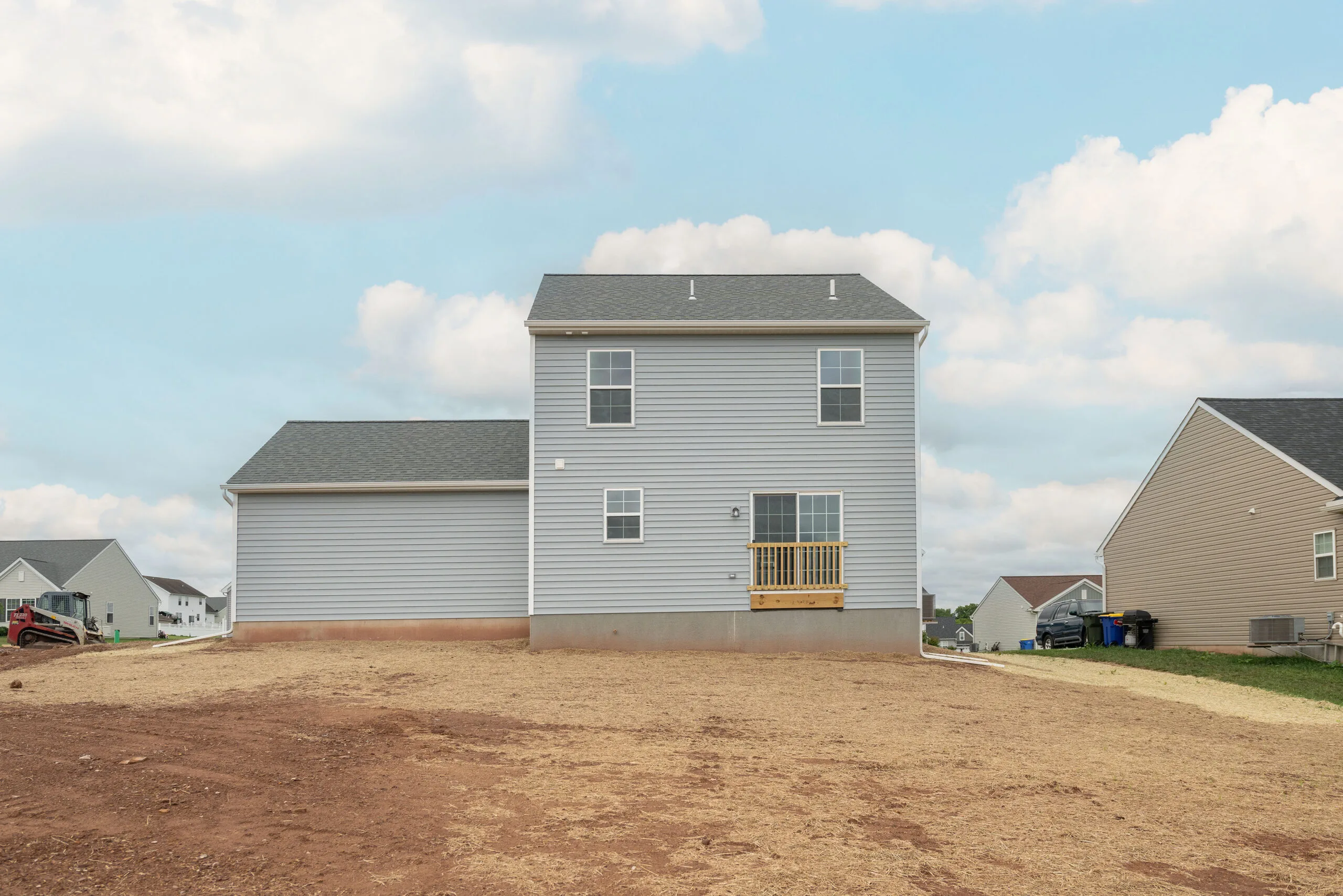 A new two-story house with gray siding and a small back deck sits on a bare dirt yard in a suburban neighborhood under a partly cloudy sky. Other houses are visible nearby.