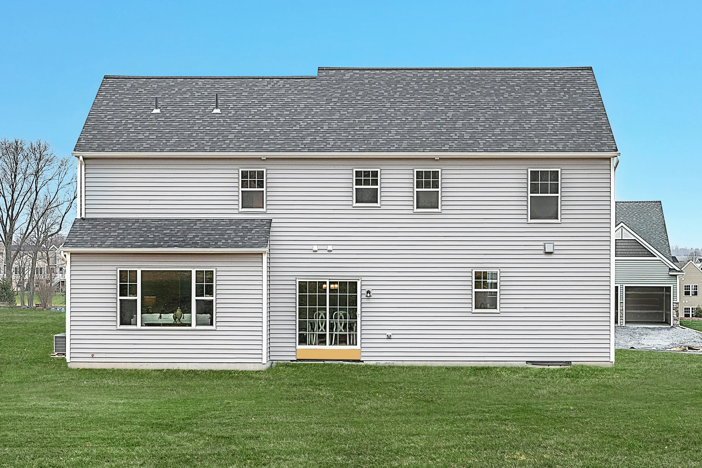 The back view of a two-story house with light-colored siding, several windows, a yellow door leading to a small patio, and a large green lawn under a clear blue sky.