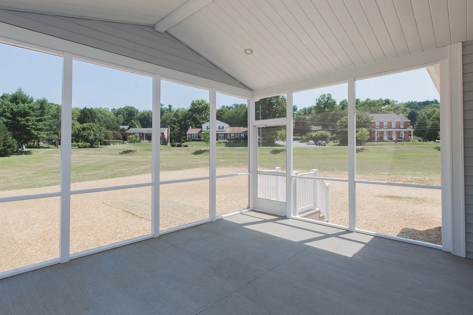 A screened-in porch with a white ceiling and supports, concrete floor, and views of a grassy yard with trees and houses in the distance under a clear blue sky.