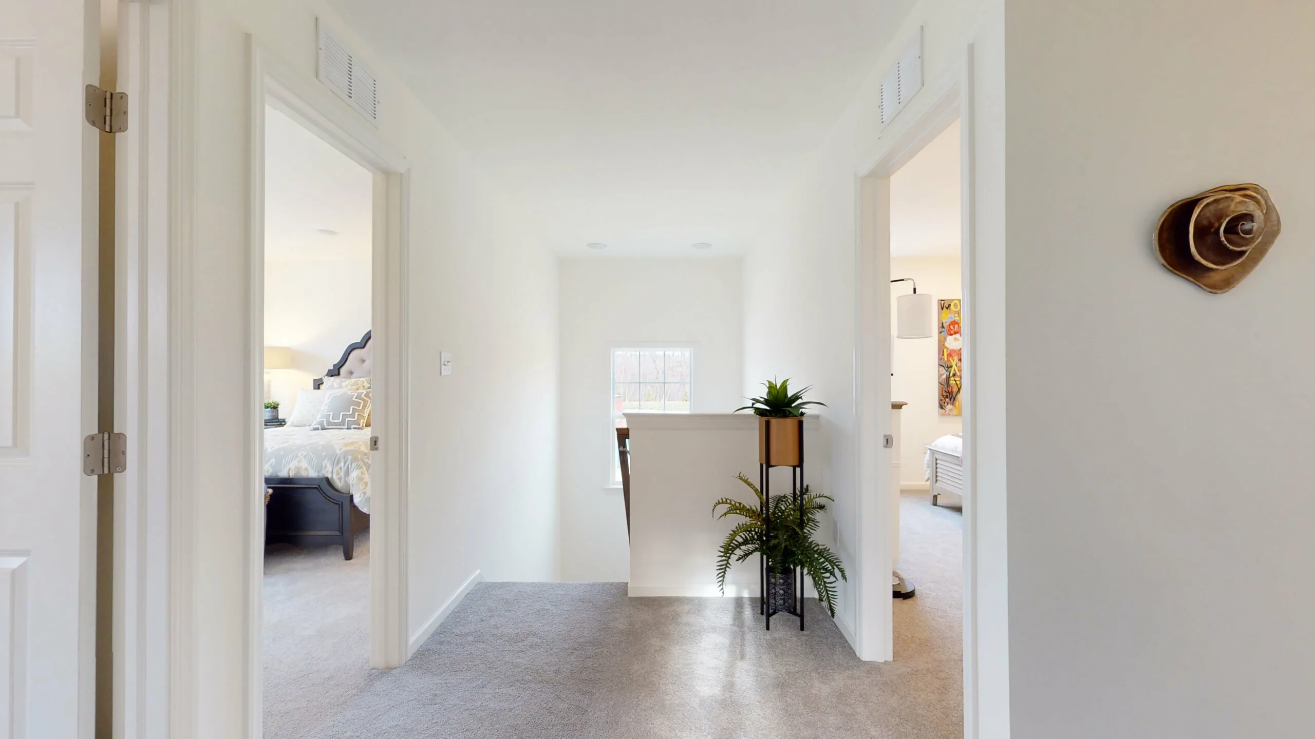 A bright hallway with light-colored walls and carpet, two bedrooms on each side, and a small window at the end. Two potted plants on a stand are placed near the stair railing in the center.