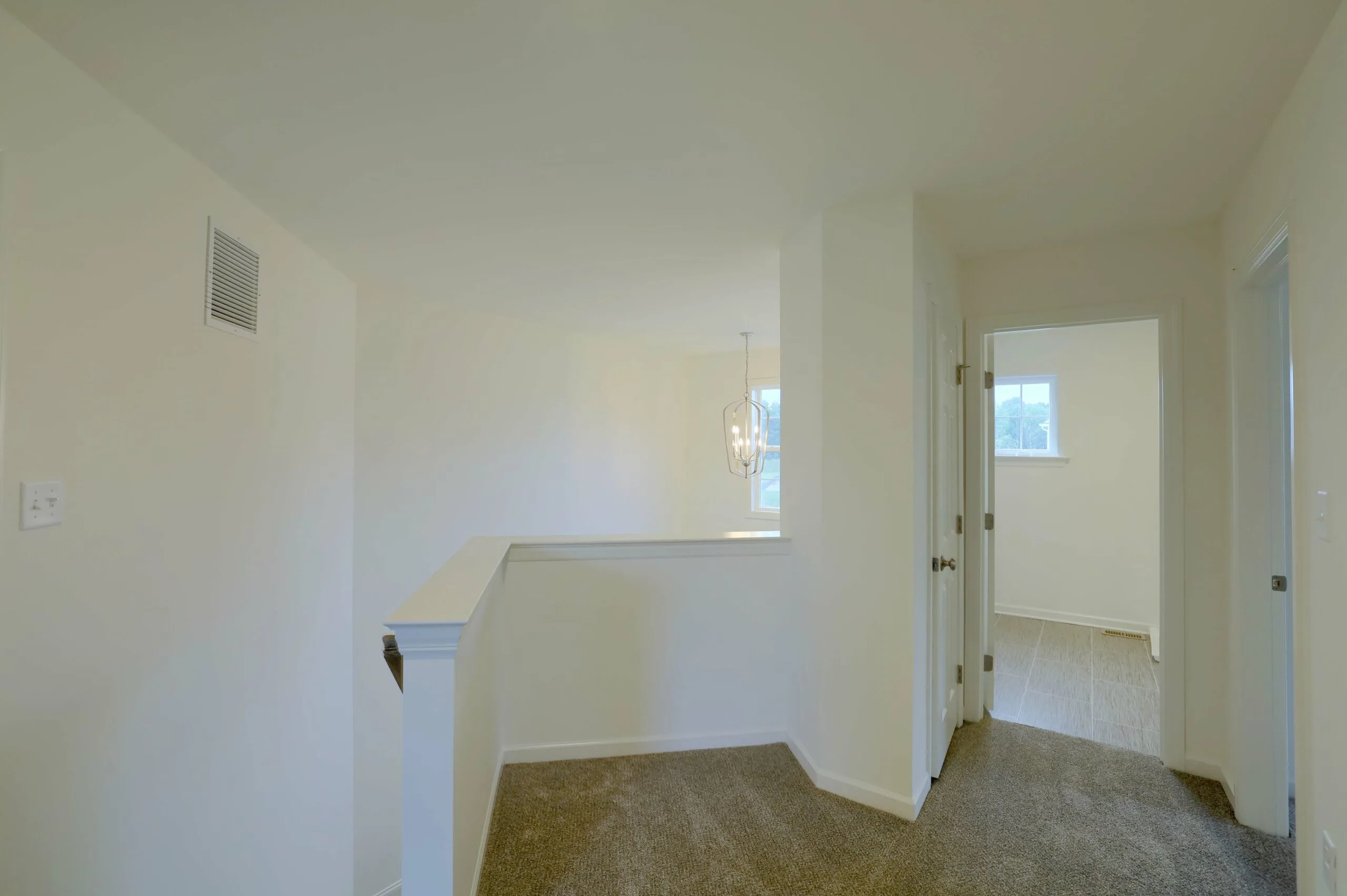 Second-floor landing in a house with beige carpet, white walls, a short railing overlooking a dining area with a modern light fixture, and doorways leading to other rooms with natural light.
