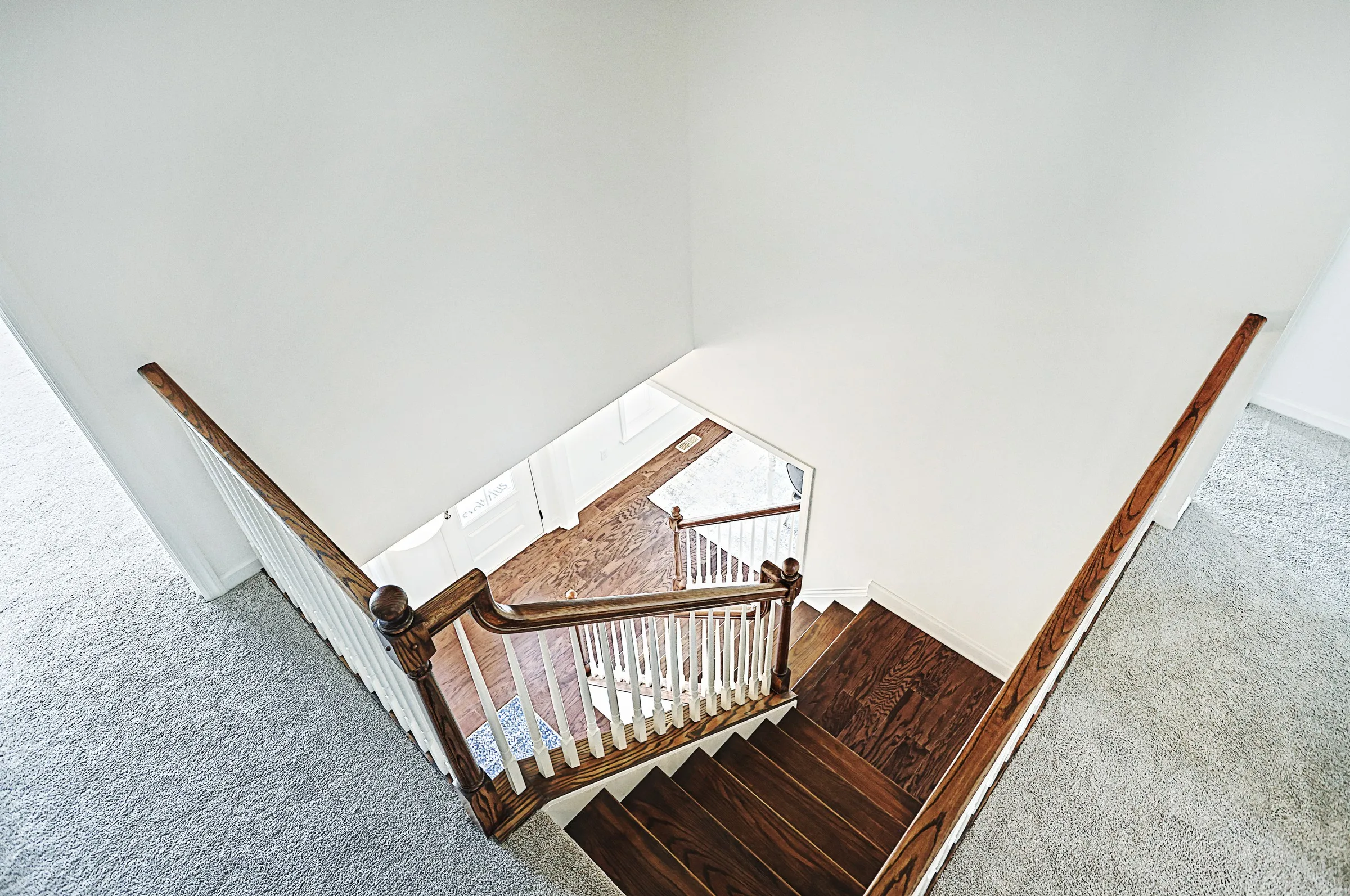View from above of a wooden staircase with white railings in a home, leading down to a landing and front entryway. The stairs are surrounded by light-colored carpet and walls.