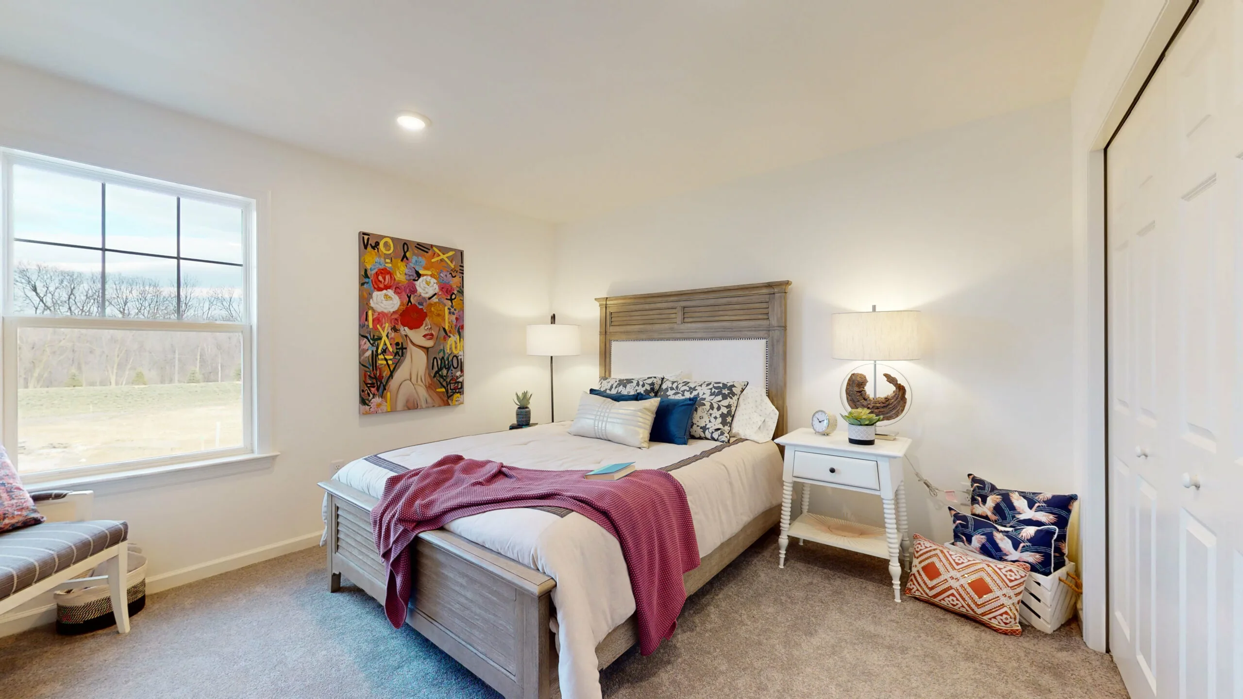 A cozy bedroom with a wooden bed, white and blue bedding, a red throw, two bedside tables with lamps, a colorful abstract painting, a window with a view of trees, and a basket with cushions on a carpeted floor.