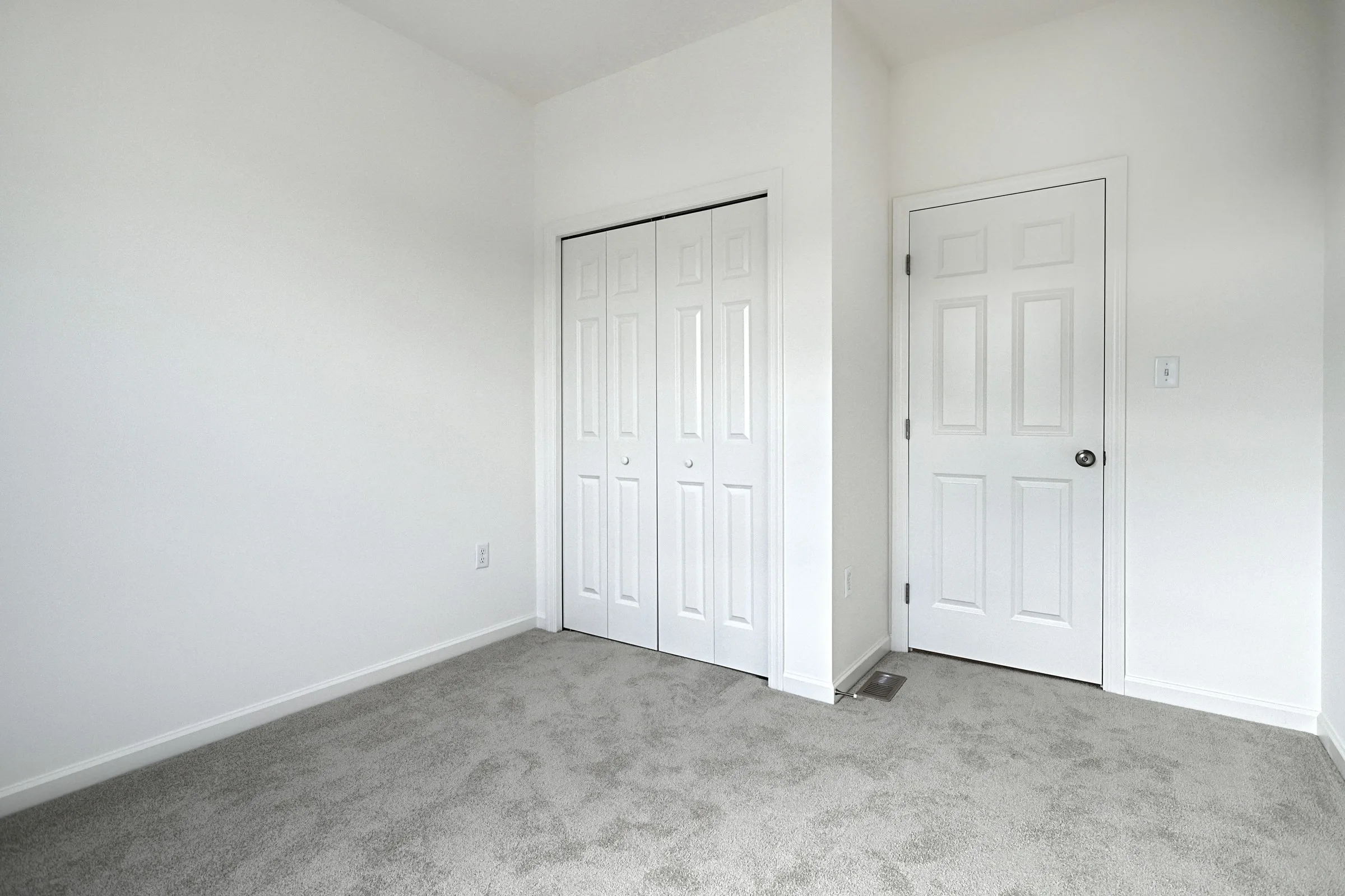 Empty room with light gray carpet, white walls, and two white doors—one closet door with double panels and one standard door. There is a floor vent near the door and an electrical outlet on the wall.