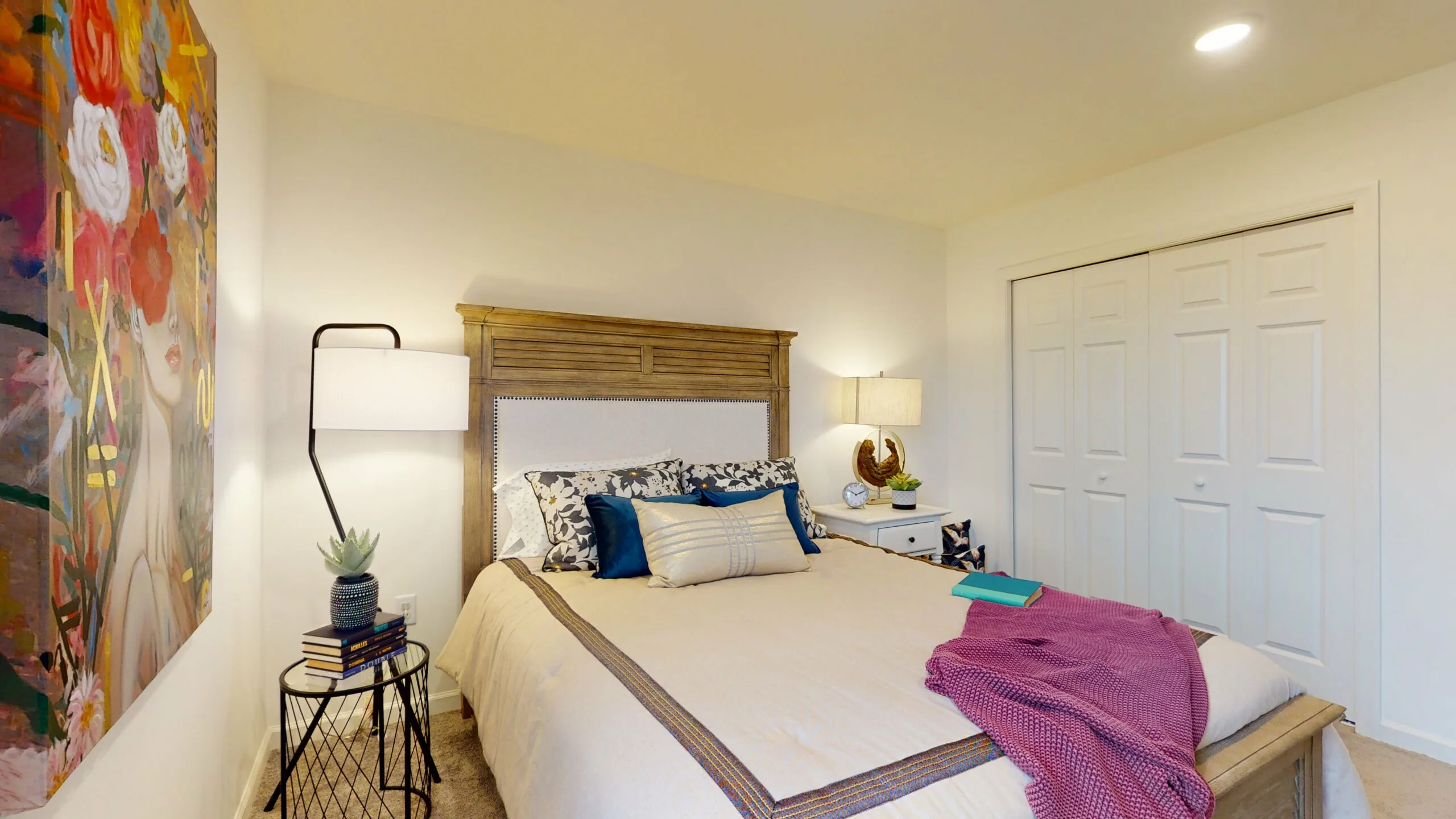 A cozy bedroom with a wooden bed, cream bedding, patterned pillows, a purple throw, and books. There is a colorful painting on the wall, white double closet doors, and two lamps on either side of the bed.