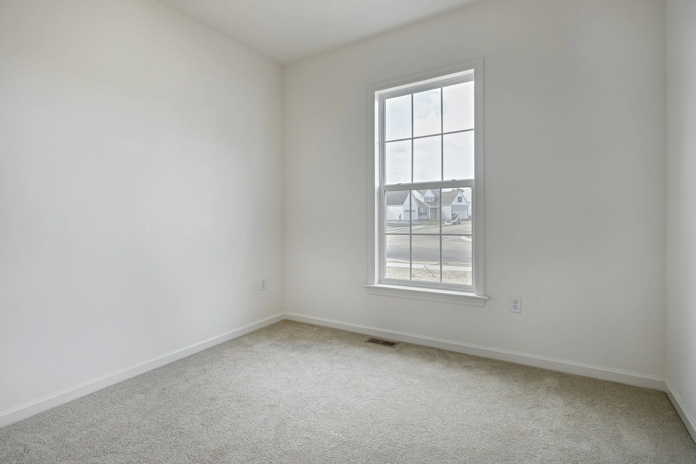 A small, empty room with white walls, light gray carpet, and a large window letting in natural light, showing houses and a street outside.