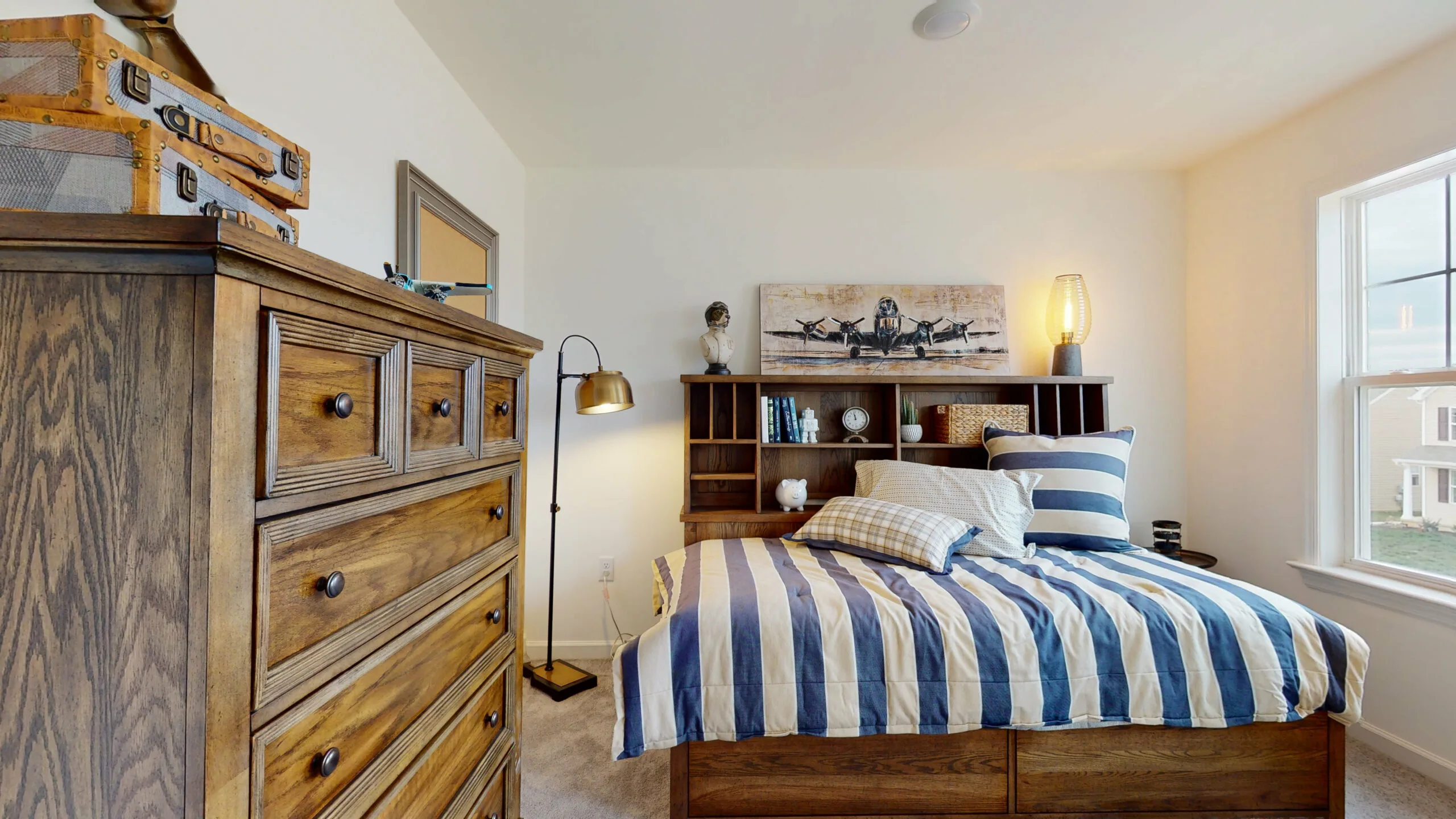 A cozy bedroom with a wooden dresser, a bed with blue and white striped bedding, decorative pillows, a shelf with books and ornaments, a floor lamp, and a window letting in natural light.