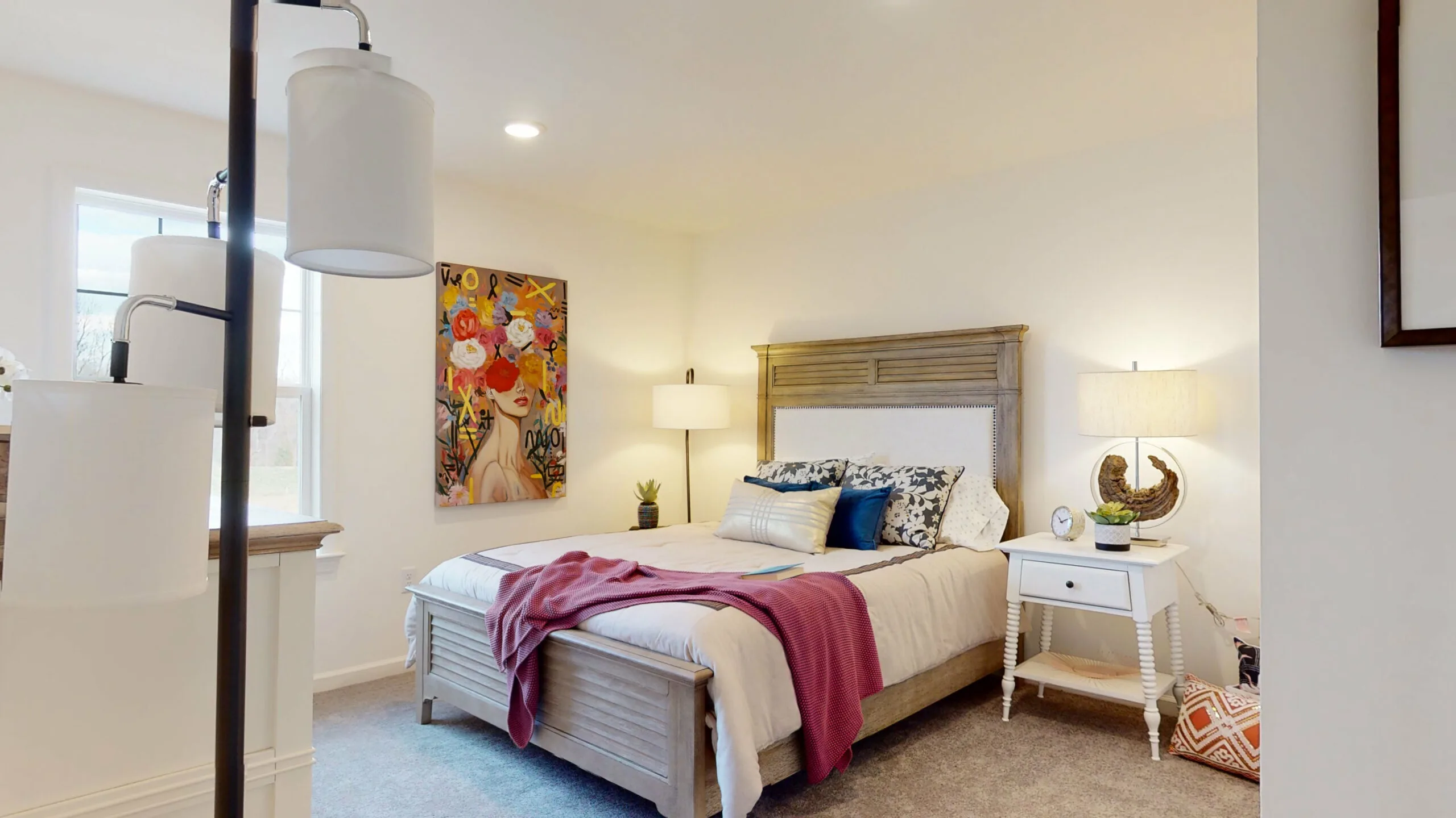 A cozy bedroom with a wooden bed, white and blue pillows, a pink throw blanket, nightstands with lamps, and a colorful floral painting on the wall. The room is softly lit and has light-colored carpeting.