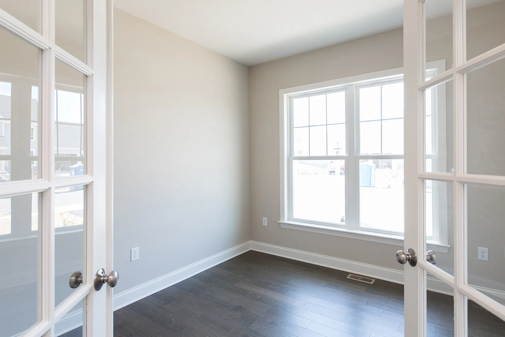 A bright, empty room with beige walls, dark wood floors, large white-framed window, and open double glass doors, letting in natural light.