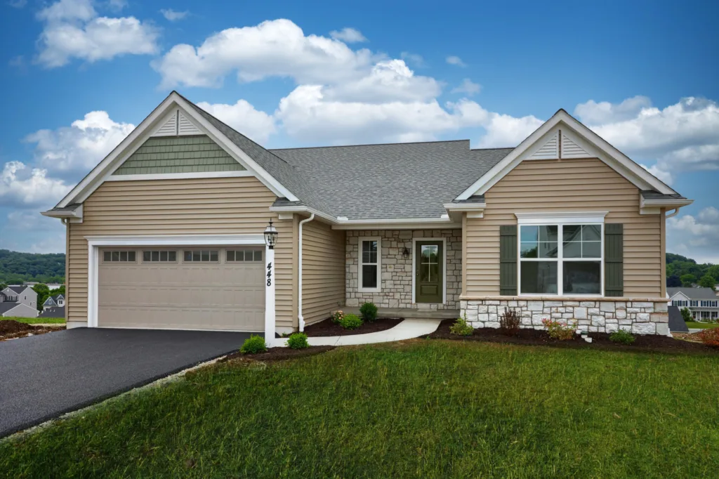 Single-story American house with tan siding, gray roof, two-car garage, green shutters, and a front porch. The partially landscaped lawn sits on a gentle hill in Summergrove under a blue sky filled with clouds.
