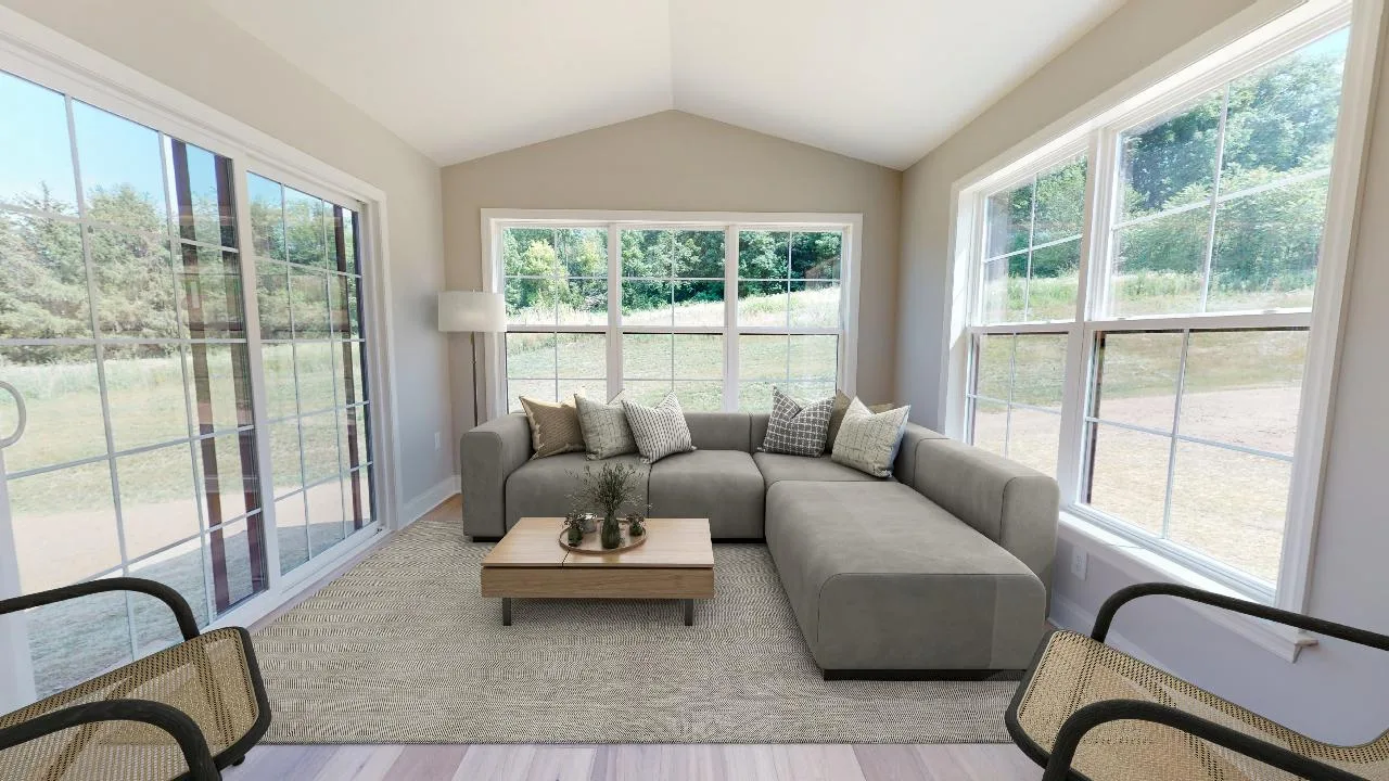 A bright sunroom with large windows, a gray sectional sofa with cushions, a wooden coffee table, two chairs, and a floor lamp on a neutral rug. Trees and sky are visible outside.