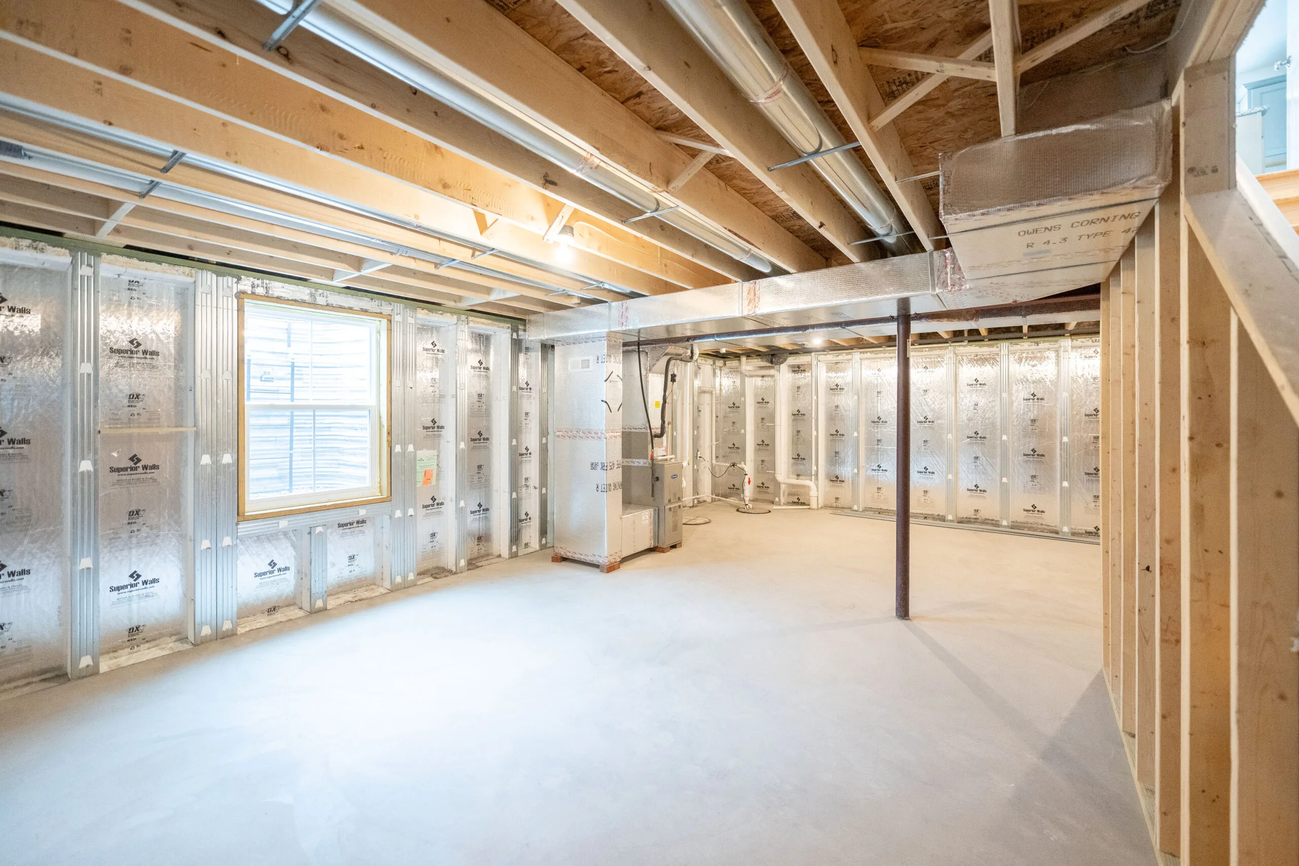 Unfinished basement with exposed wooden beams, pipes, and ductwork on the ceiling. The walls are lined with insulation and a large window lets in natural light. The concrete floor is bare and open.