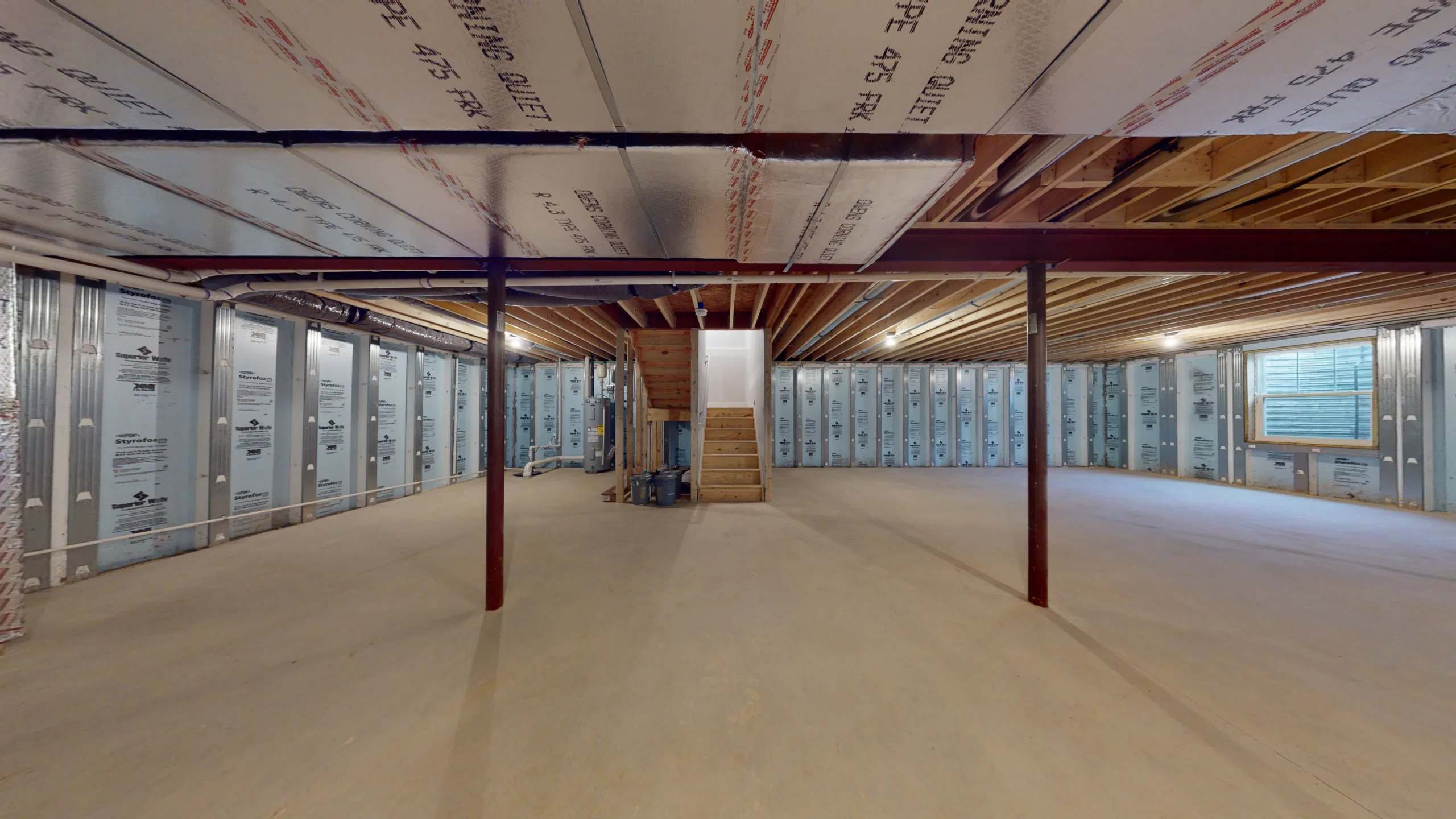 Unfinished basement with exposed beams, insulation on walls and ceiling, concrete floor, support columns, and a wooden staircase leading upstairs. Construction materials are visible near the stairs.
