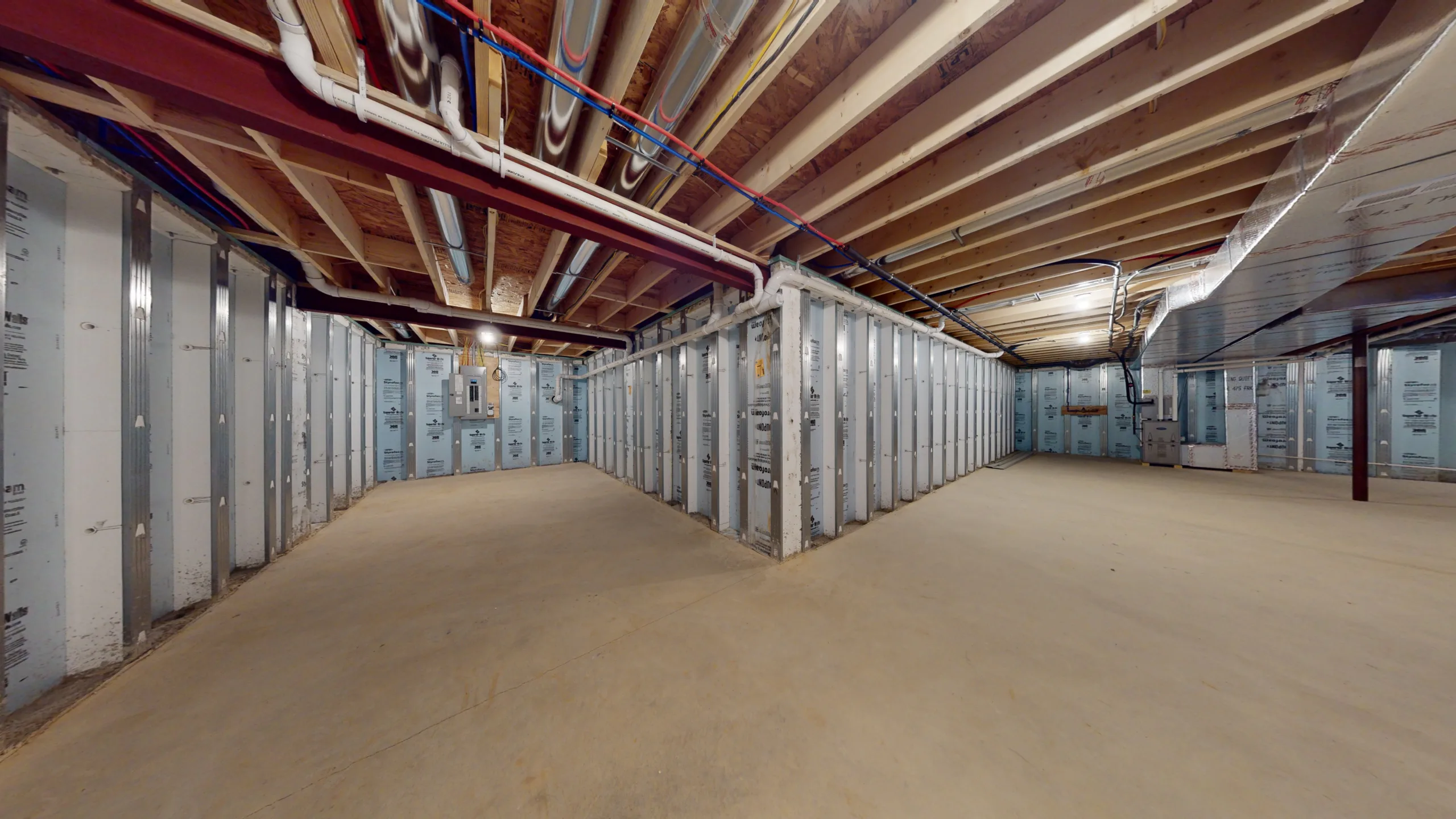A large, unfinished basement with exposed ceiling beams, pipes, and insulated walls, featuring a clean concrete floor and bright overhead lighting.
