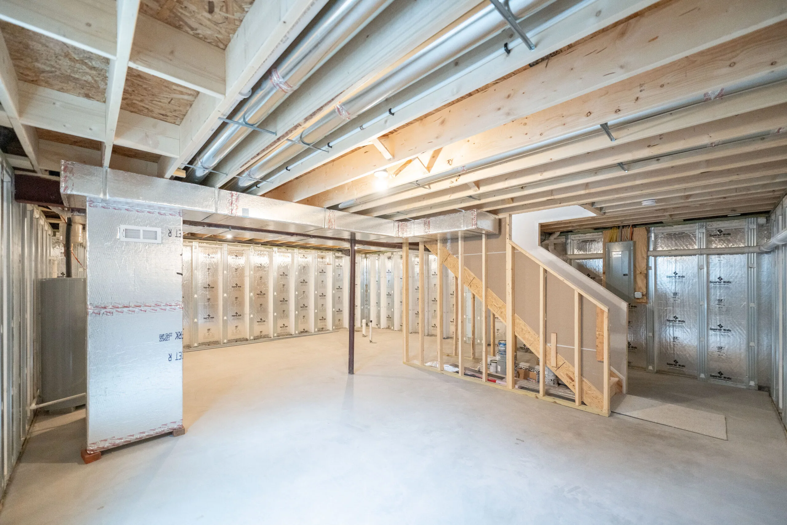 Unfinished basement with exposed wooden ceiling beams, metal air ducts, and insulated walls. Wooden stairs lead up, and the floor is bare concrete. The space is well-lit by ceiling lights.