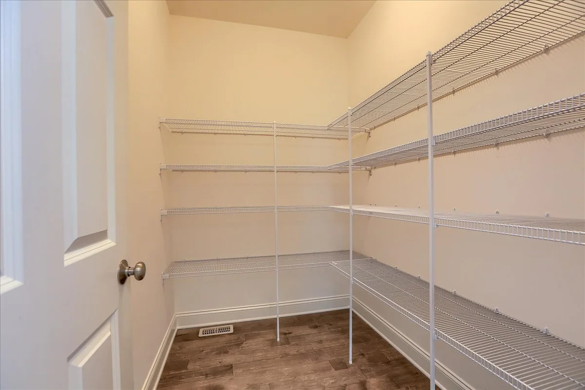 A large, empty pantry with beige walls, wood-style flooring, and several rows of white metal wire shelves on both sides and at the back. A white door with a silver knob is partially visible on the left.