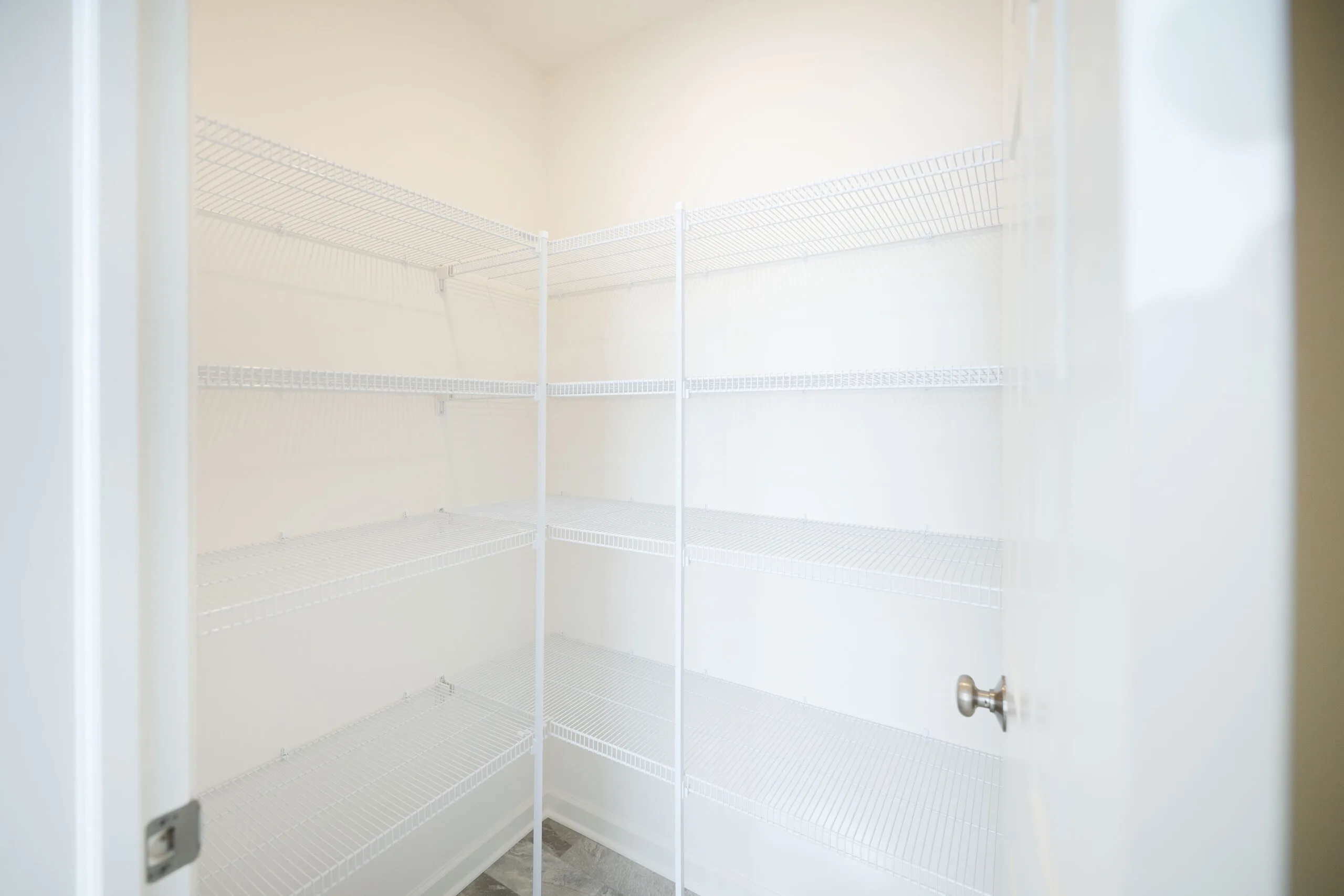 Empty walk-in pantry with white wire shelving on two walls, beige walls, and a gray tile floor. The door is open, revealing the clean and spacious interior.
