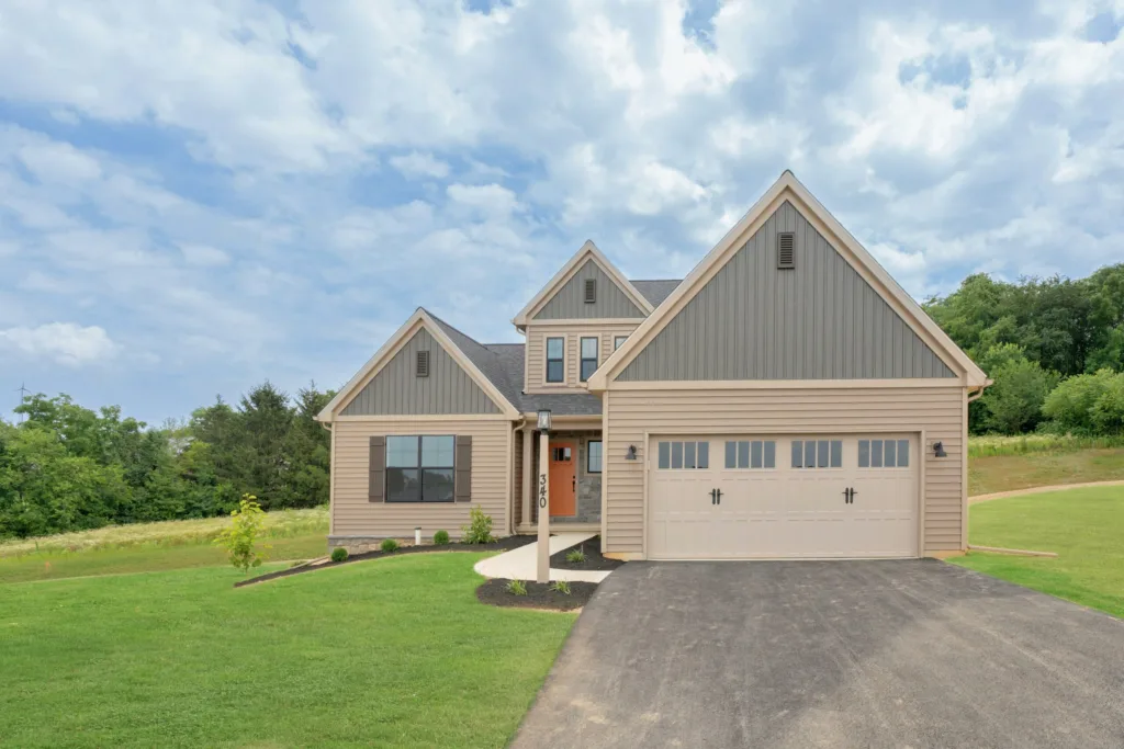 A modern beige and gray two-story suburban house with a double garage, manicured lawn, young trees, and a paved driveway under a partly cloudy sky.