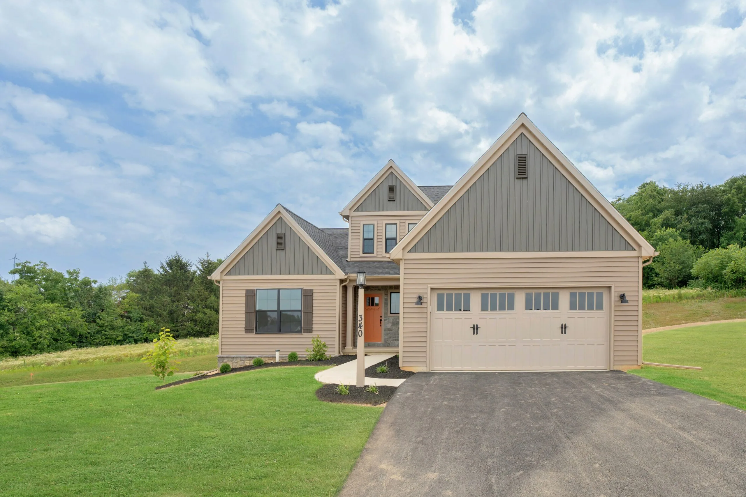 A modern beige and gray two-story suburban house with a double garage, manicured lawn, young trees, and a paved driveway under a partly cloudy sky.