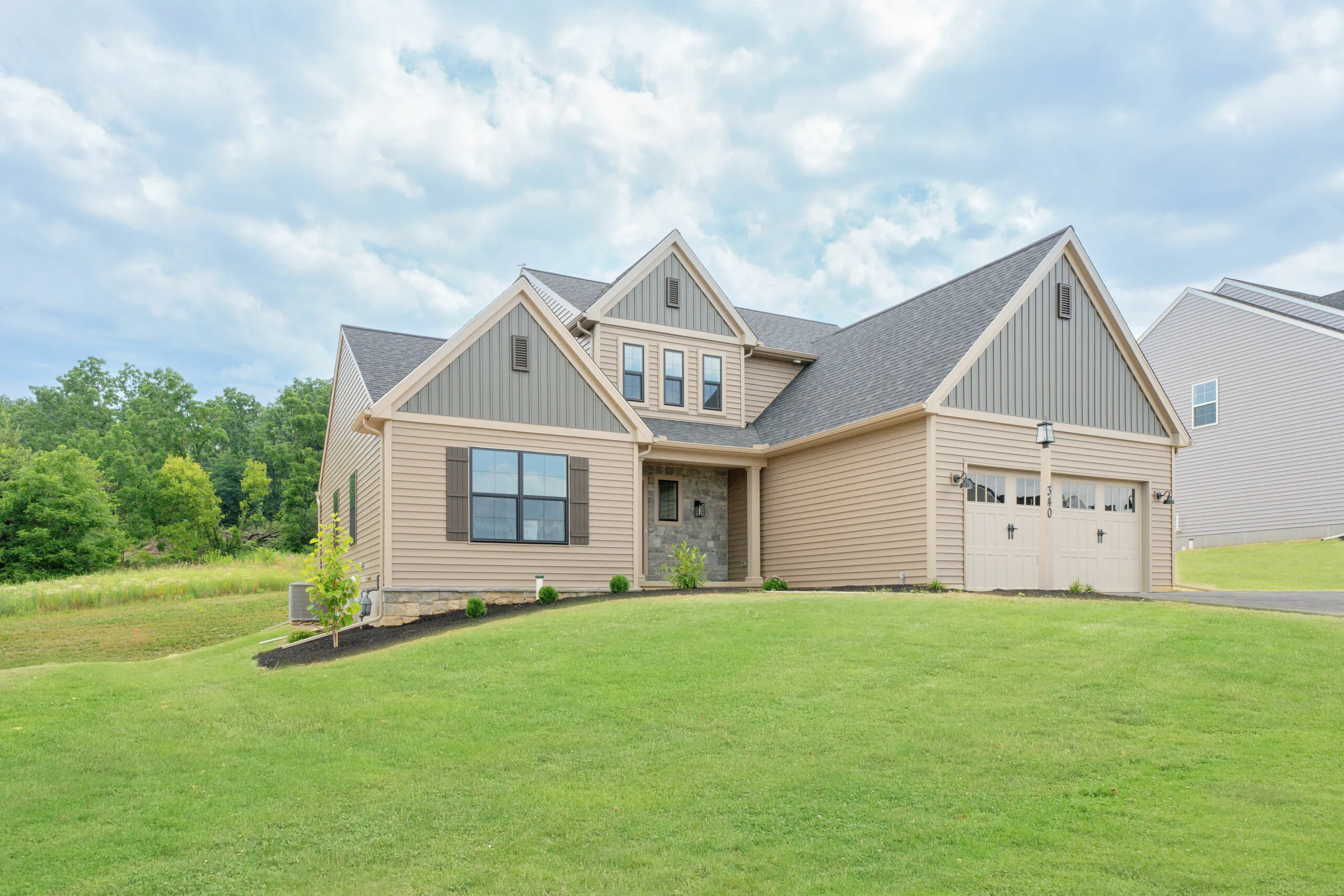 A modern beige house with gray accents, large windows, and a double garage sits on a sloped green lawn with landscaped shrubs under a partly cloudy sky.