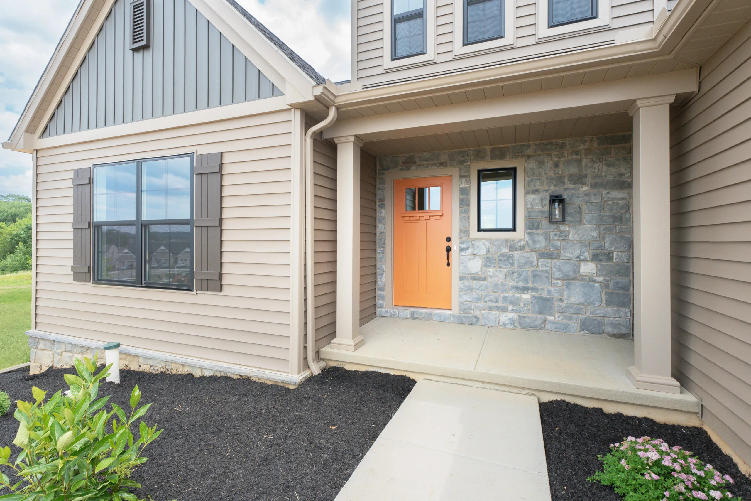 Front entrance of a modern house with beige siding, stone facade, an orange front door, a small window, columns, and a concrete walkway, surrounded by mulch and green plants.