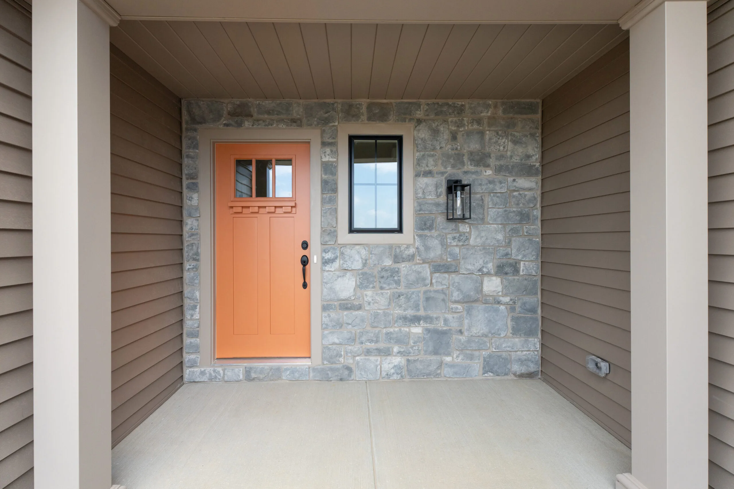 A covered entryway with tan siding and stone wall features an orange front door, a narrow rectangular window, and a modern black wall lantern. The floor is smooth concrete.