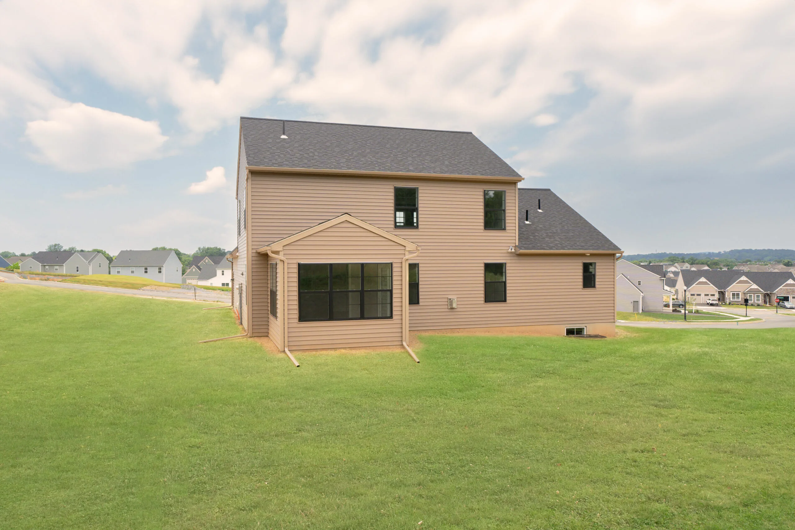 A tan two-story house with black-framed windows sits on a large, grassy lawn under a partly cloudy sky. Other houses and a street are visible in the background.
