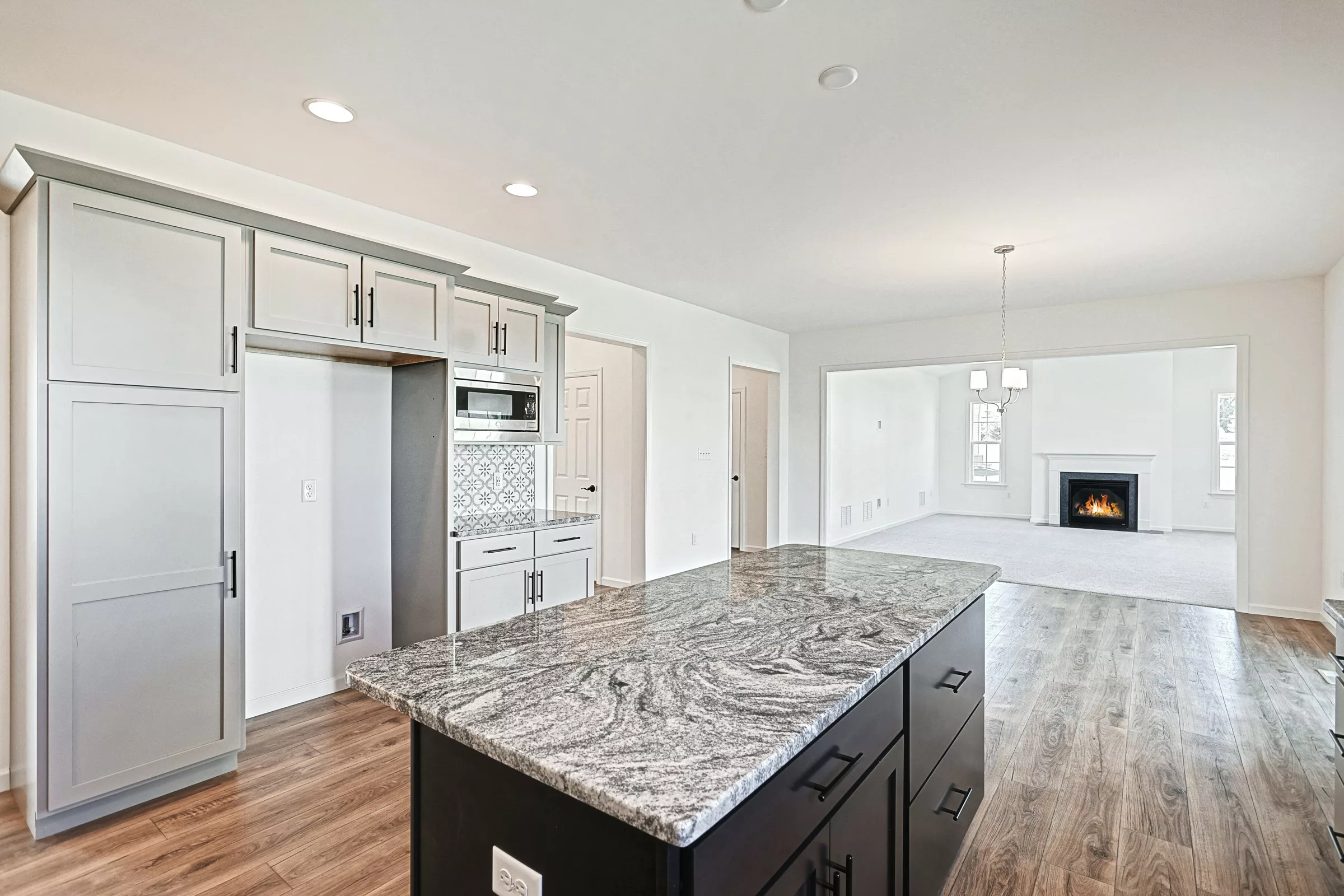Modern kitchen with gray cabinets, granite island countertop, wood floors, and stainless appliances, opening to a bright living room with carpet, windows, chandelier, and a lit fireplace in the background.