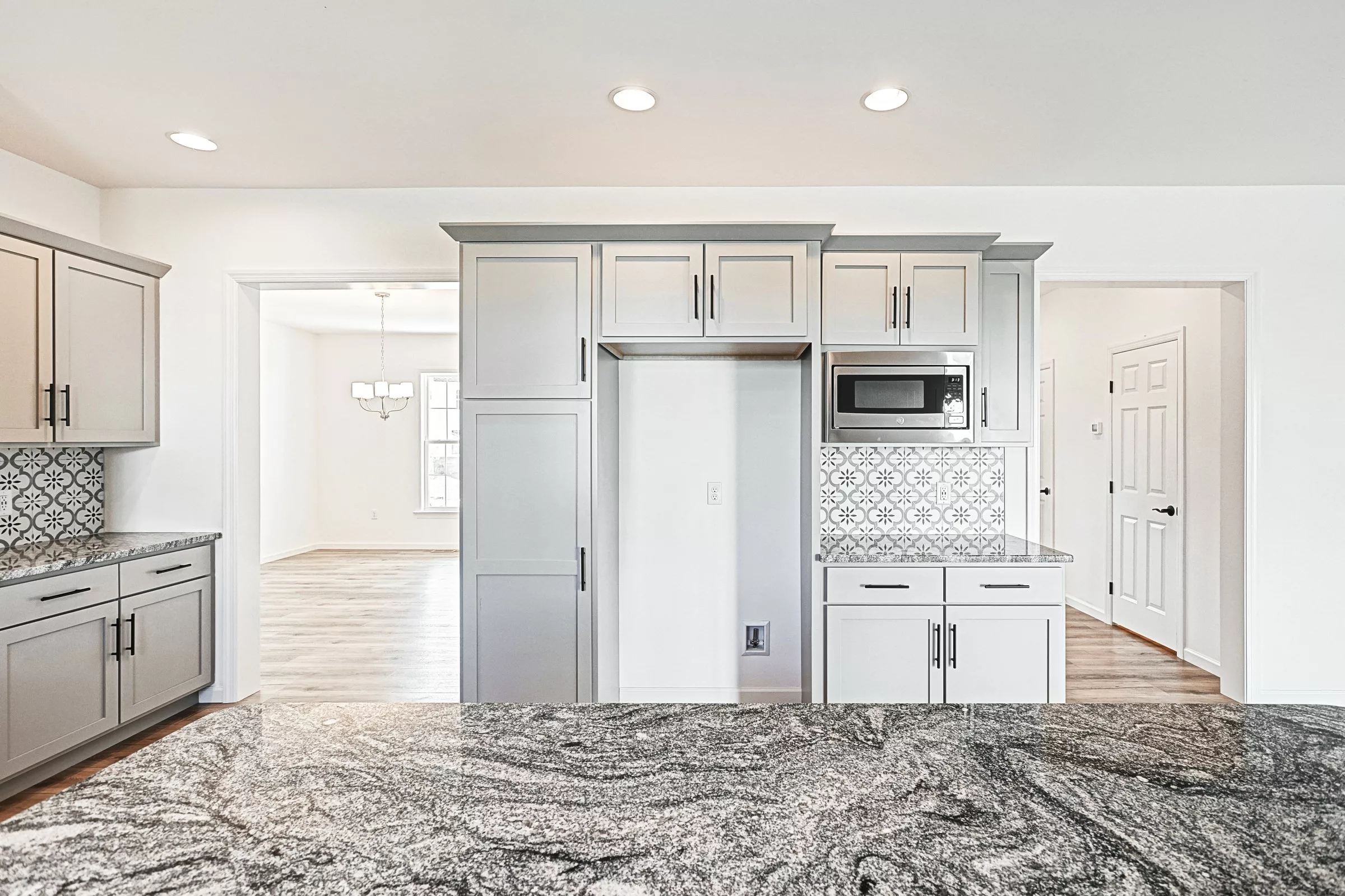 Modern kitchen with light gray cabinets, granite countertops, a built-in microwave, patterned tile backsplash, and wood flooring. An open doorway leads to a bright dining area with a window and chandelier.