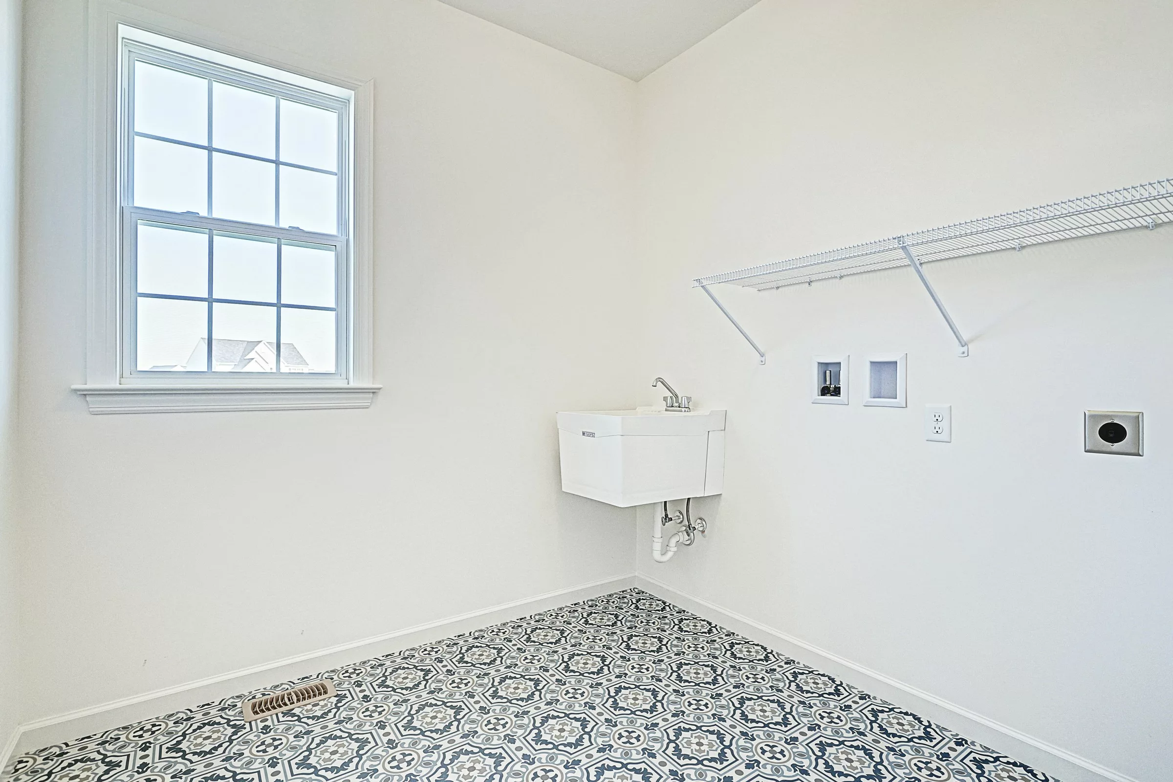 A bright laundry room with a patterned tile floor, white walls, a window, a utility sink, wall-mounted faucet, wire shelf, and outlets for a washer and dryer. The room is empty and well-lit.