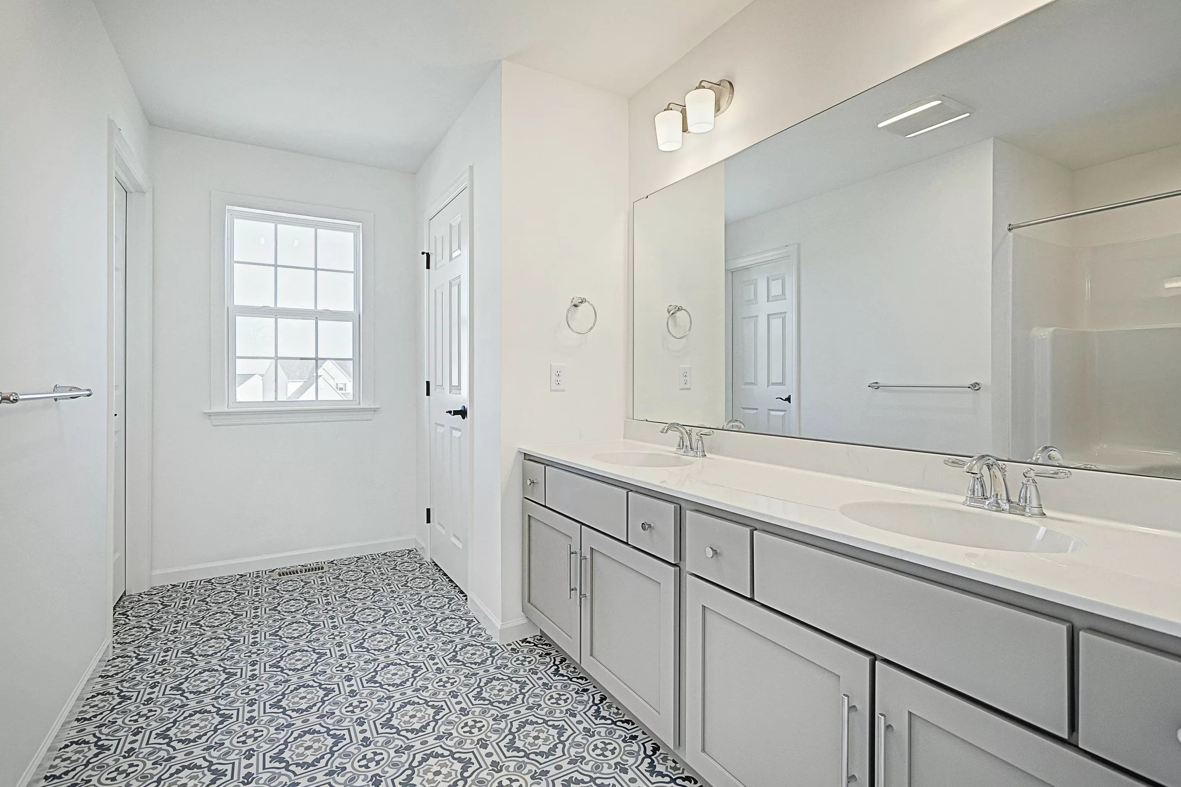 A bright, modern bathroom with patterned tile flooring, double sinks with gray cabinets, a large mirror, and a window letting in natural light.